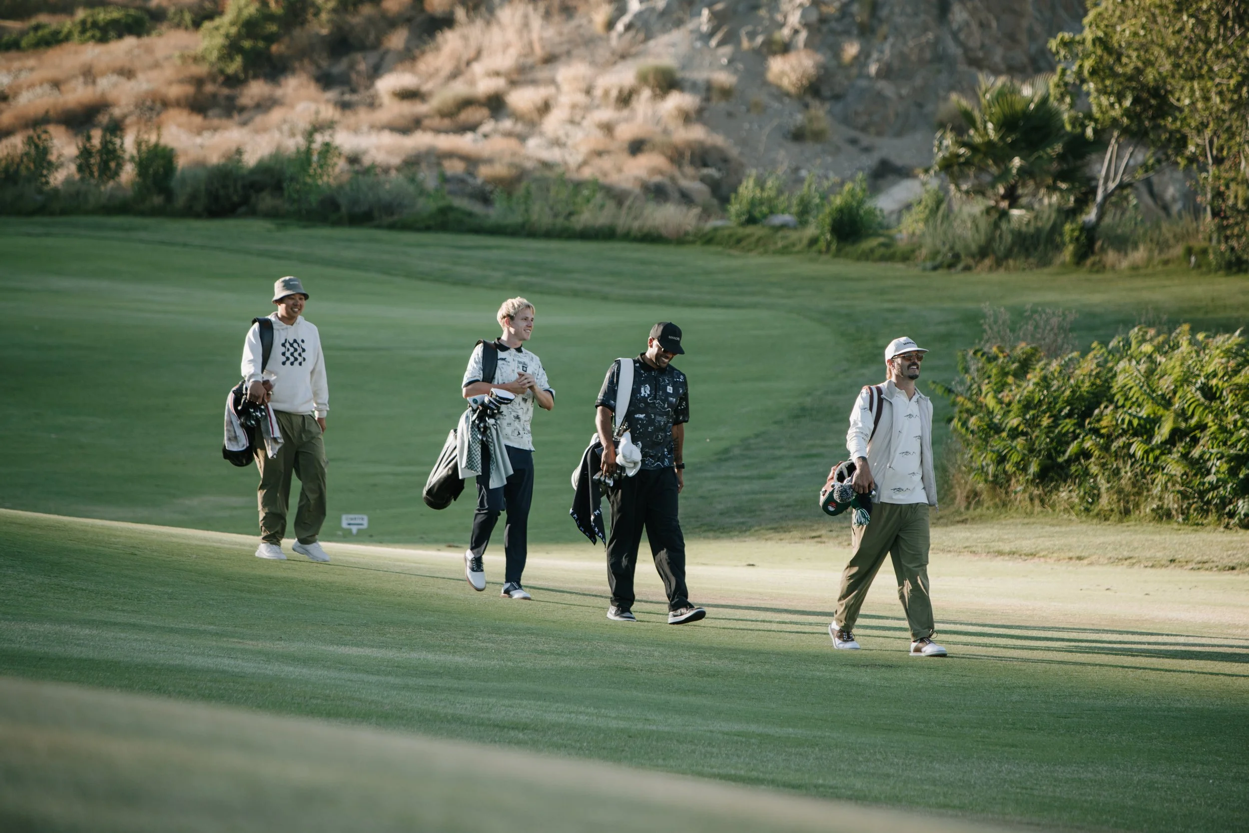 Four men walking on a golf course, each carrying golf bags and clubs, with a rocky hillside and trees in the background.