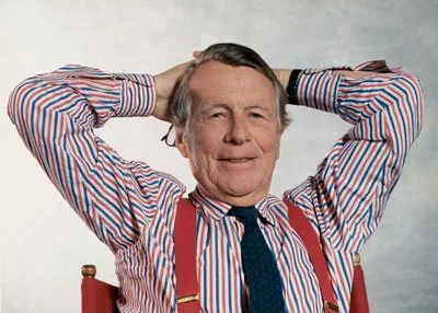 An older man with gray hair, wearing a striped shirt and suspenders, sitting with hands behind his head and smiling.