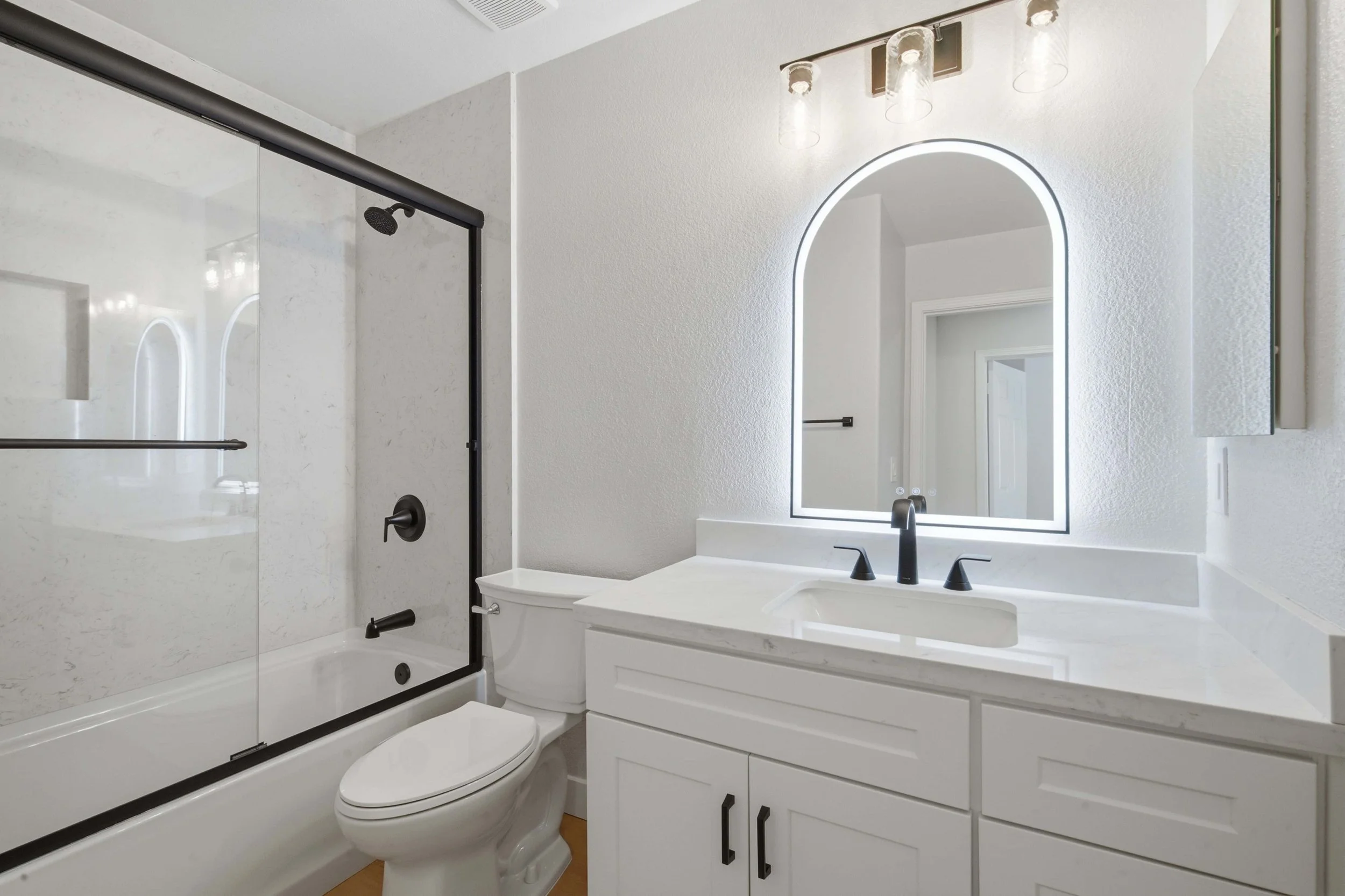 Bathroom in a renovated Bay Area condo. This room has a new tub and glass shower enclosure. There is on the right side of the photo a single vanity with white marble countertop and a three pendant light. A new toilet sits between the shower and vanit