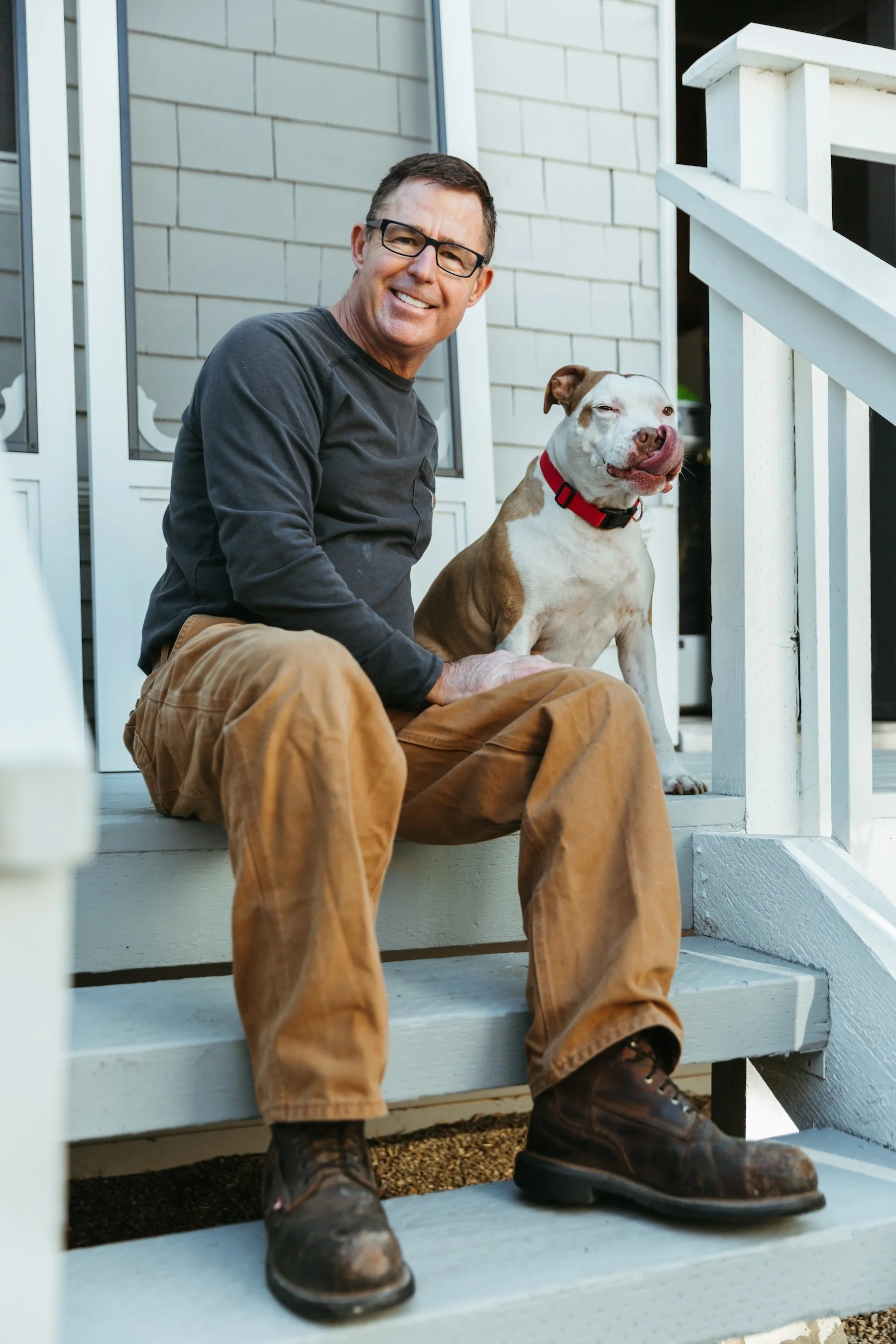 Stephen Bass, a man with glasses in a black shirt and brown pants, sits on a porch step with a white and brown dog. The dog is sitting next to him, winking. The porch has white railings and a house with gray siding in the background.