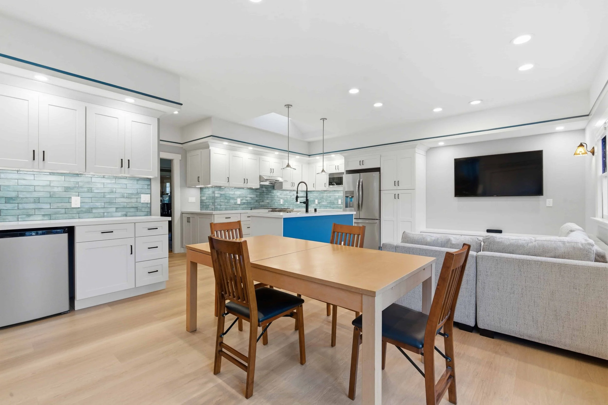 Kitchen and dining area in a remodeled home. The wood table and chairs are in the foreground with the kitchen and media area in the background.