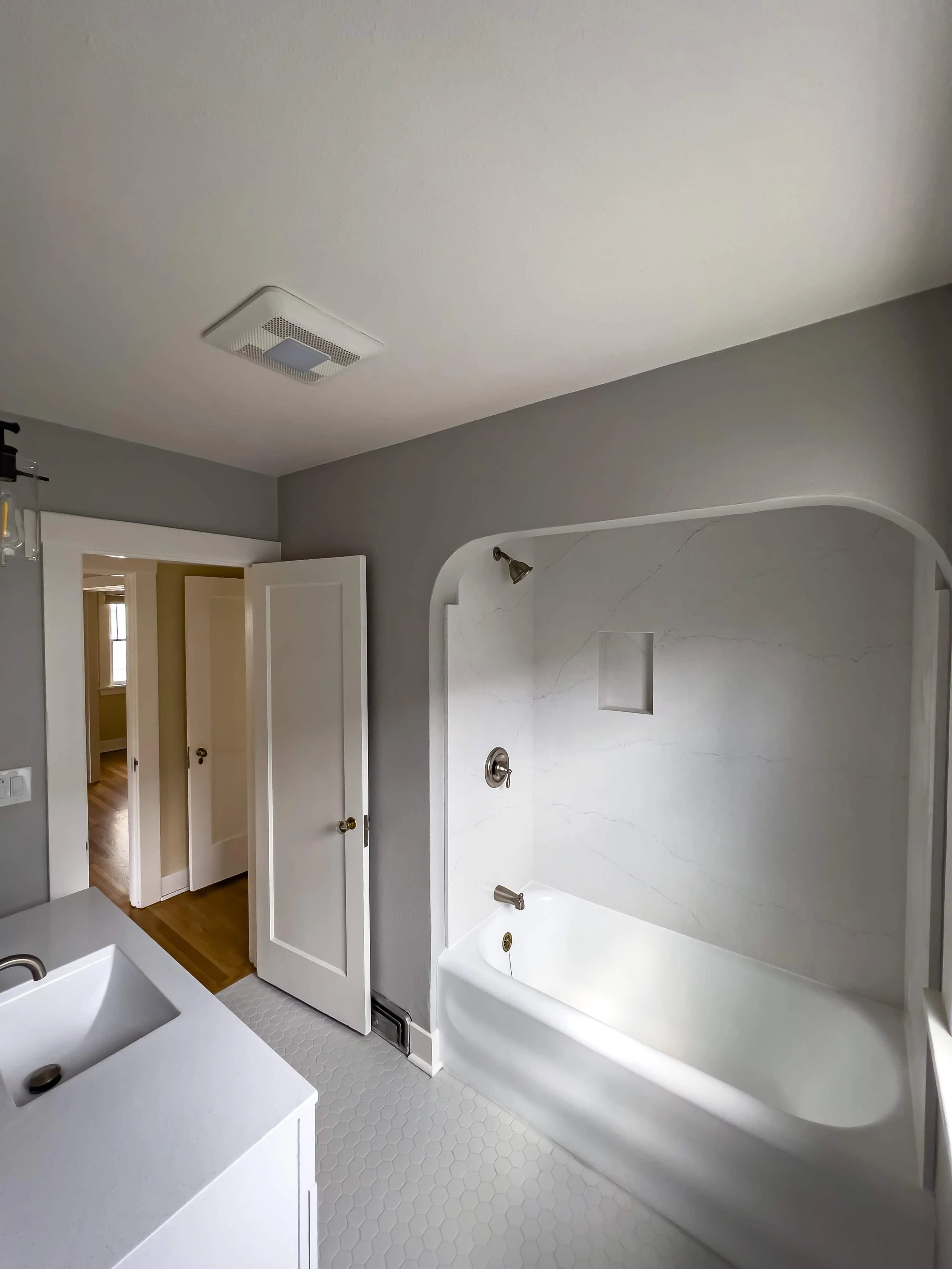 View from the far wall of renovated bathroom showing the inset tub with marble enclosure and white tile flooring.