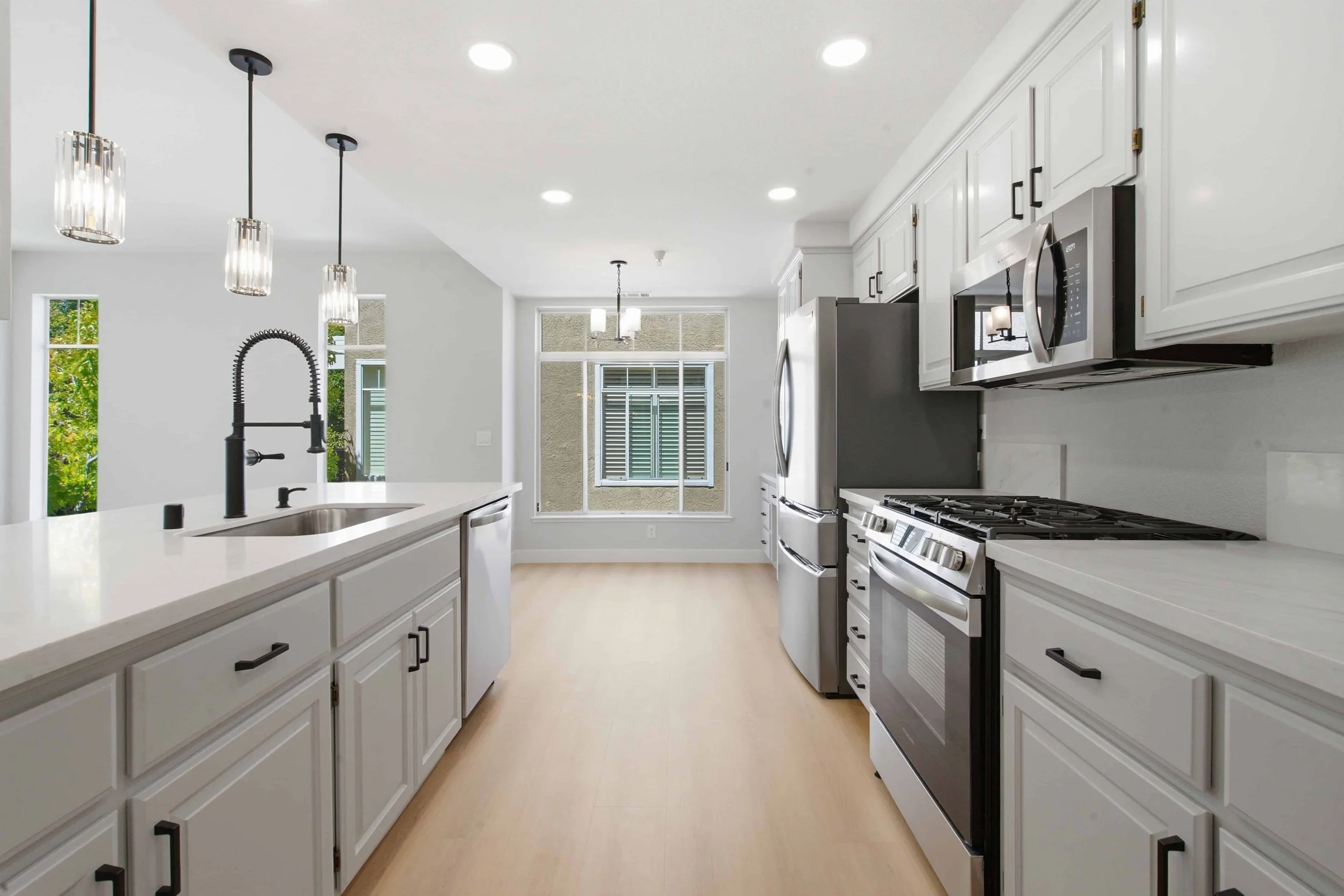 Interior long image from between a kitchen island and the back cabinets. The kitchen in this condo has all new gray cabinets, white marble countertops, hanging lighting and a wood floor. 