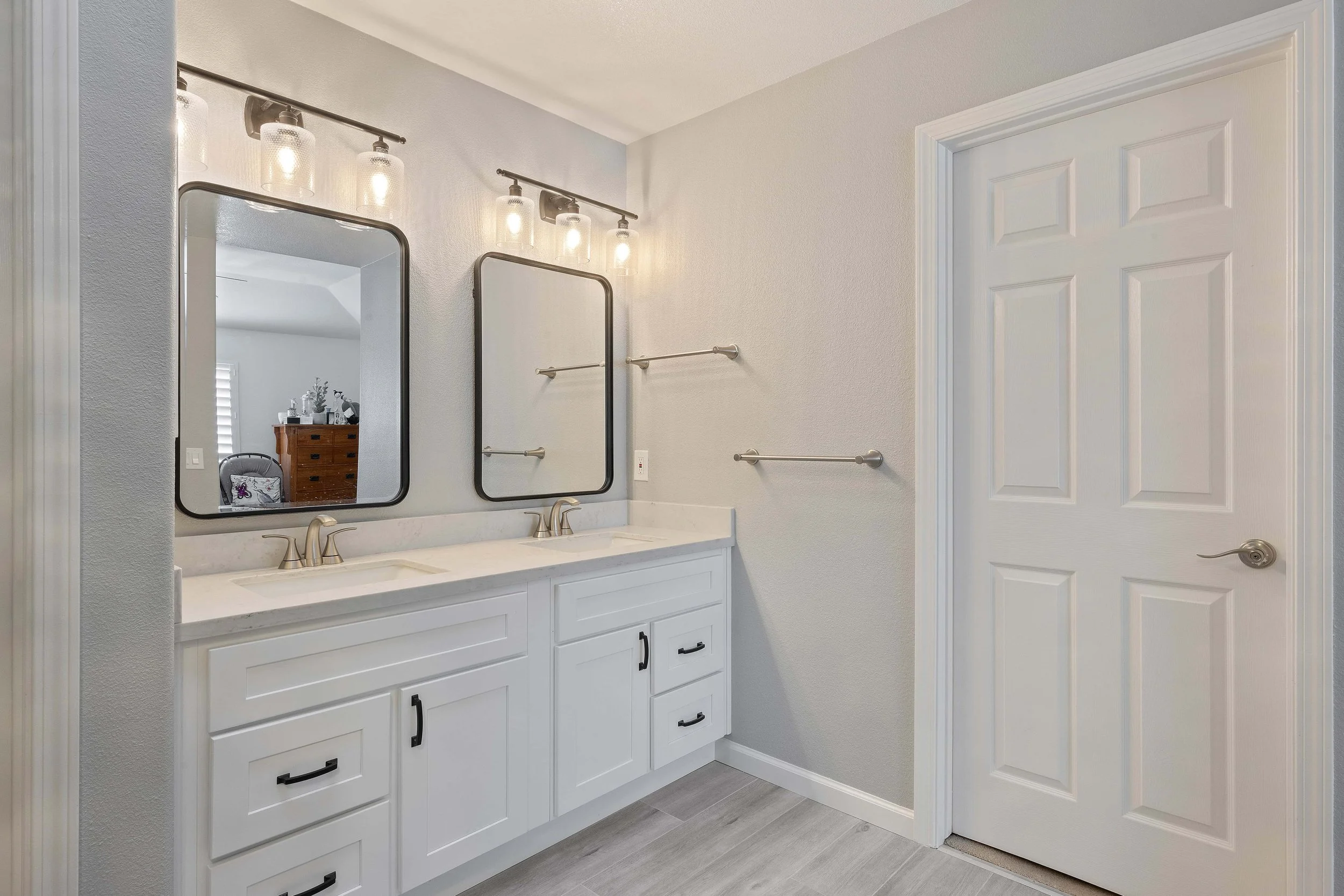 Angled view of a master bathroom vanity. The sinks and cabinets are white, as is the door. 