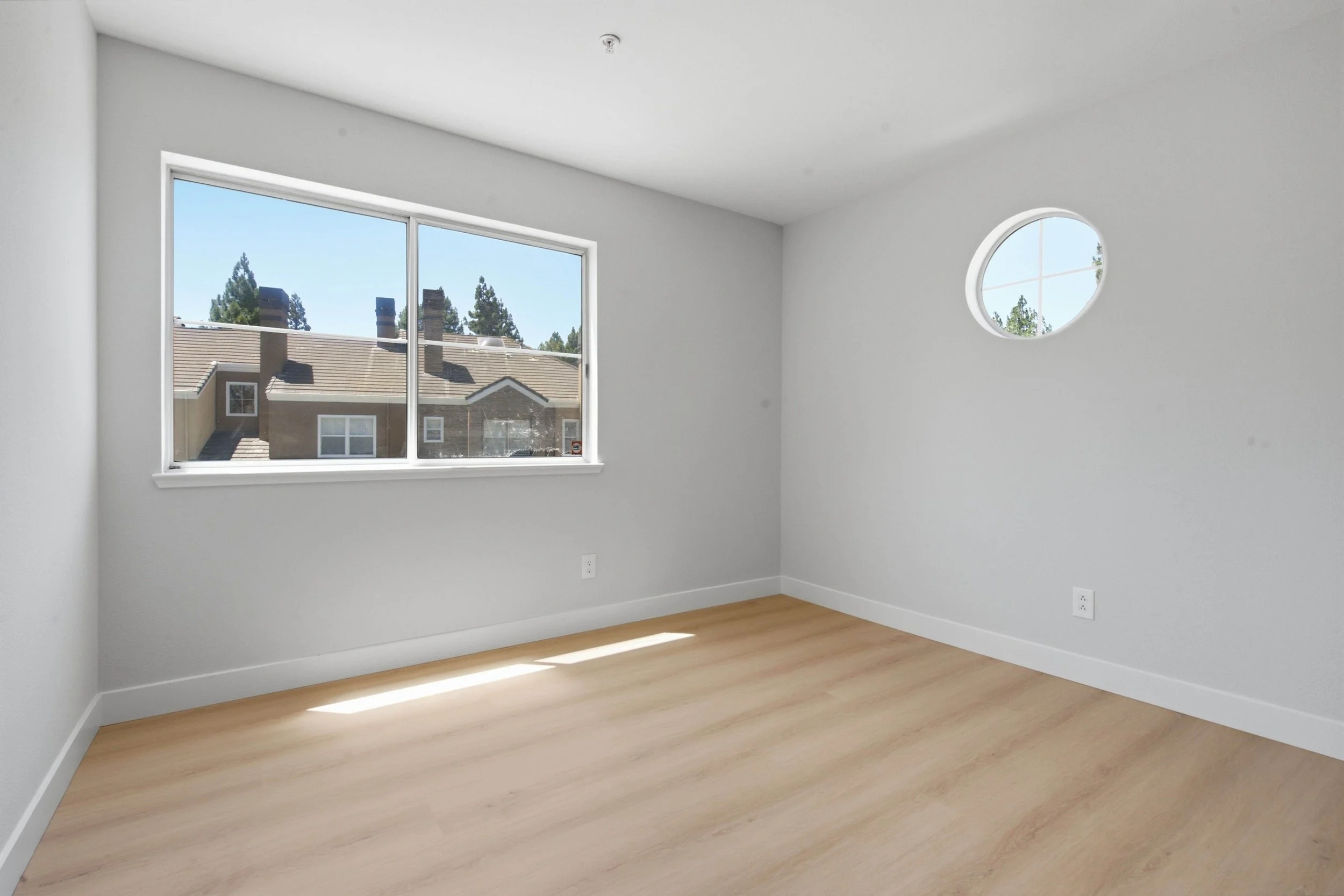 Empty bedroom in a freshly renovated California condo. The photo shows the freshly painted walls and wood floors. There is a large window overlooking other houses and on the upper right and round porthole window for privacy.