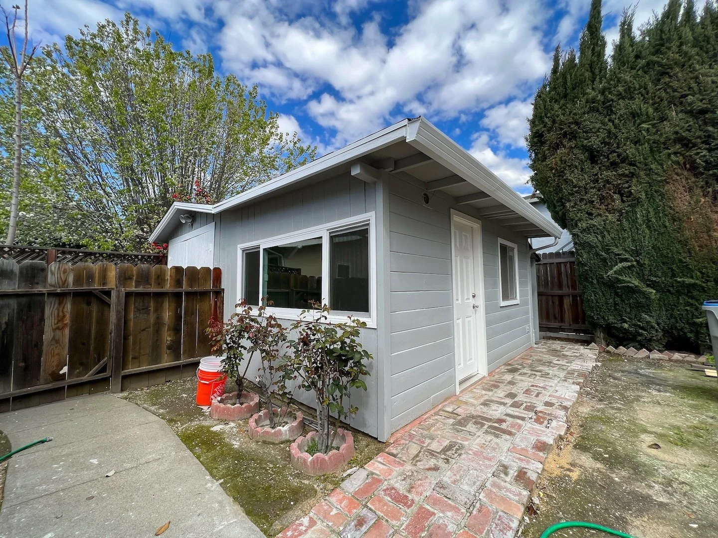 A small gray house in a backyard with a brick pathway, a wooden fence, and trees. The sky is partly cloudy.