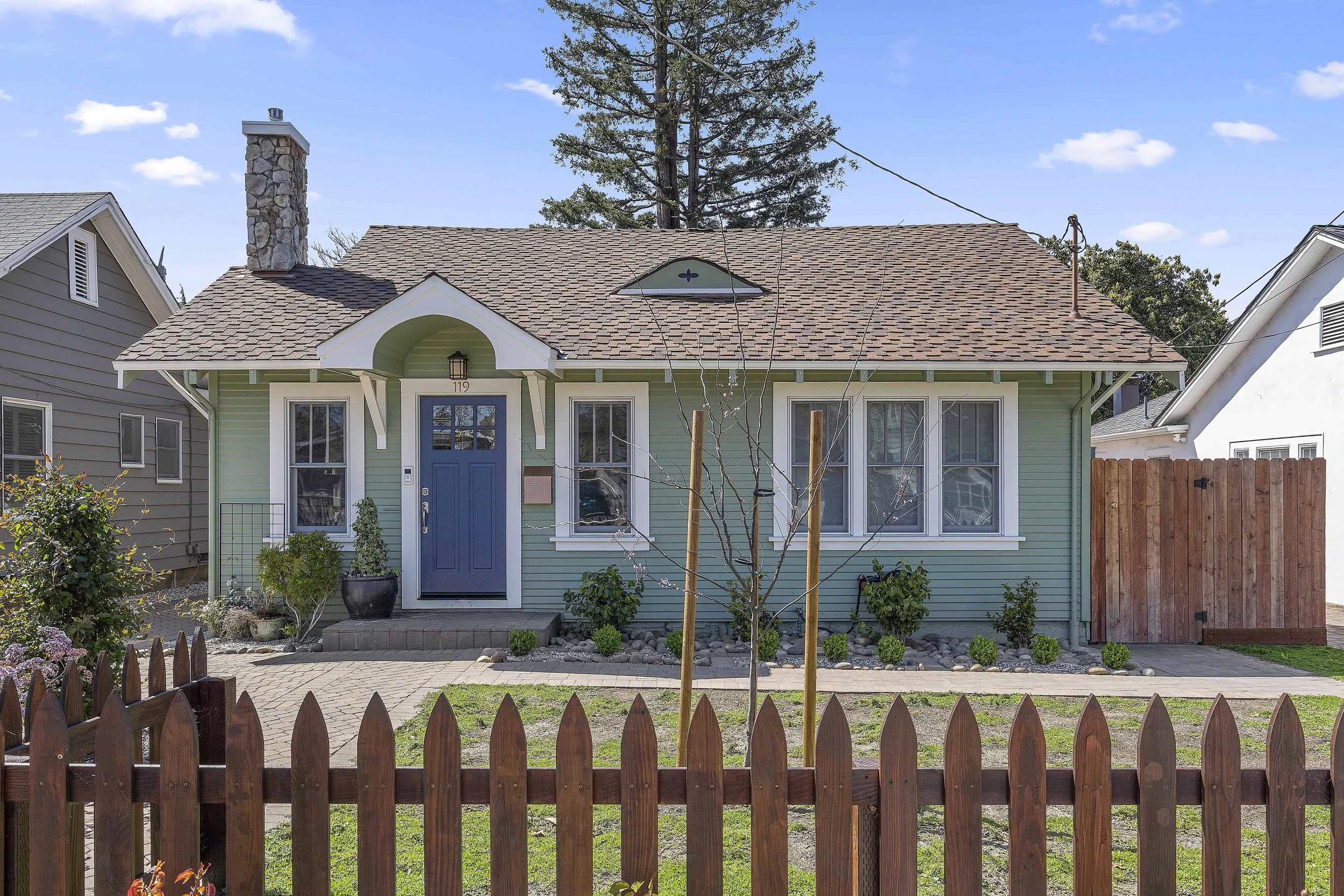 Front view of a small mint green house with a blue door and white trim, surrounded by a wooden picket fence, with a small garden and young trees in the yard, and neighboring houses visible on each side under a blue sky with some clouds.