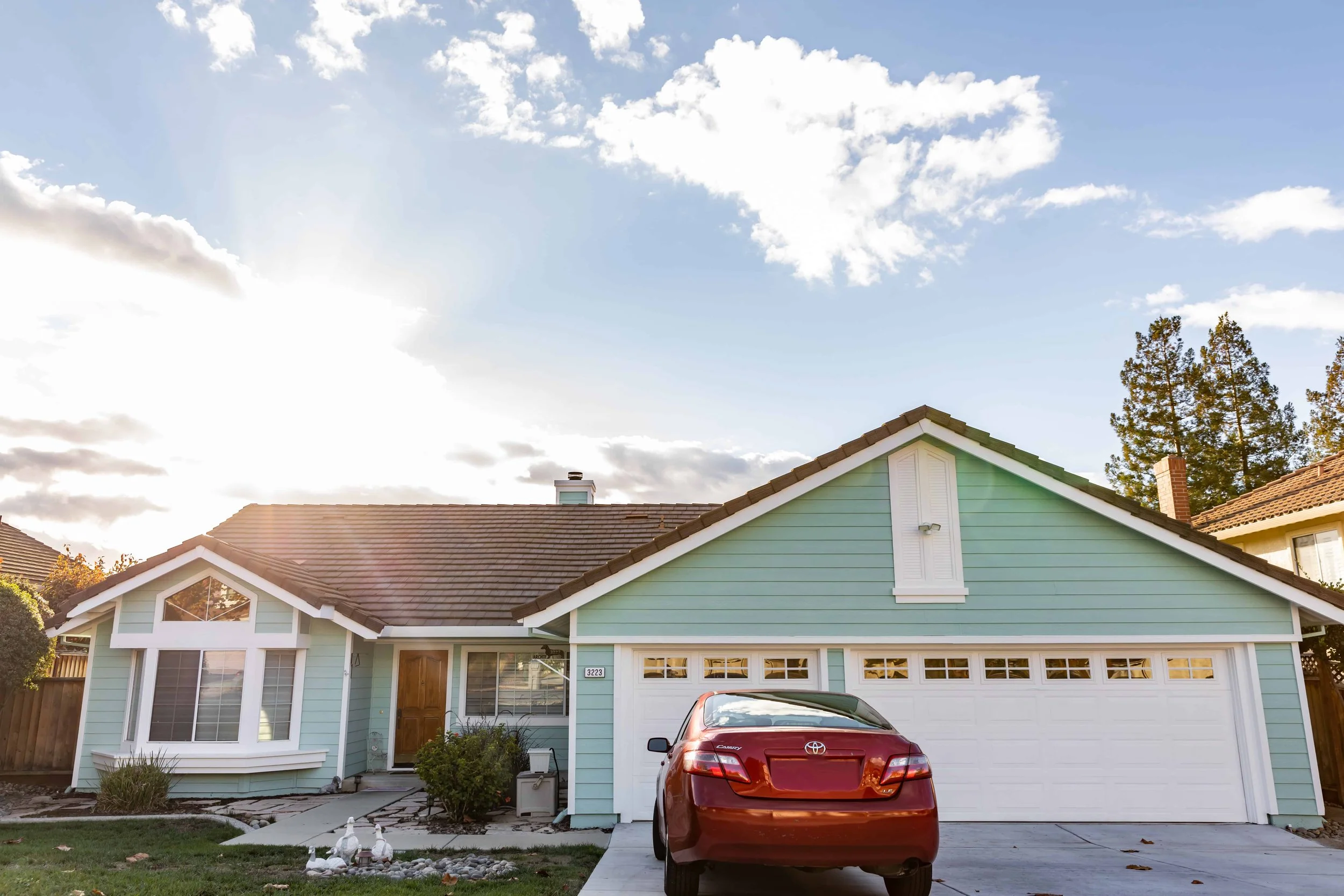 Freshly painted and horizontally sided one story sage colored house with white trim and garage doors. A red car sits in the driveway.