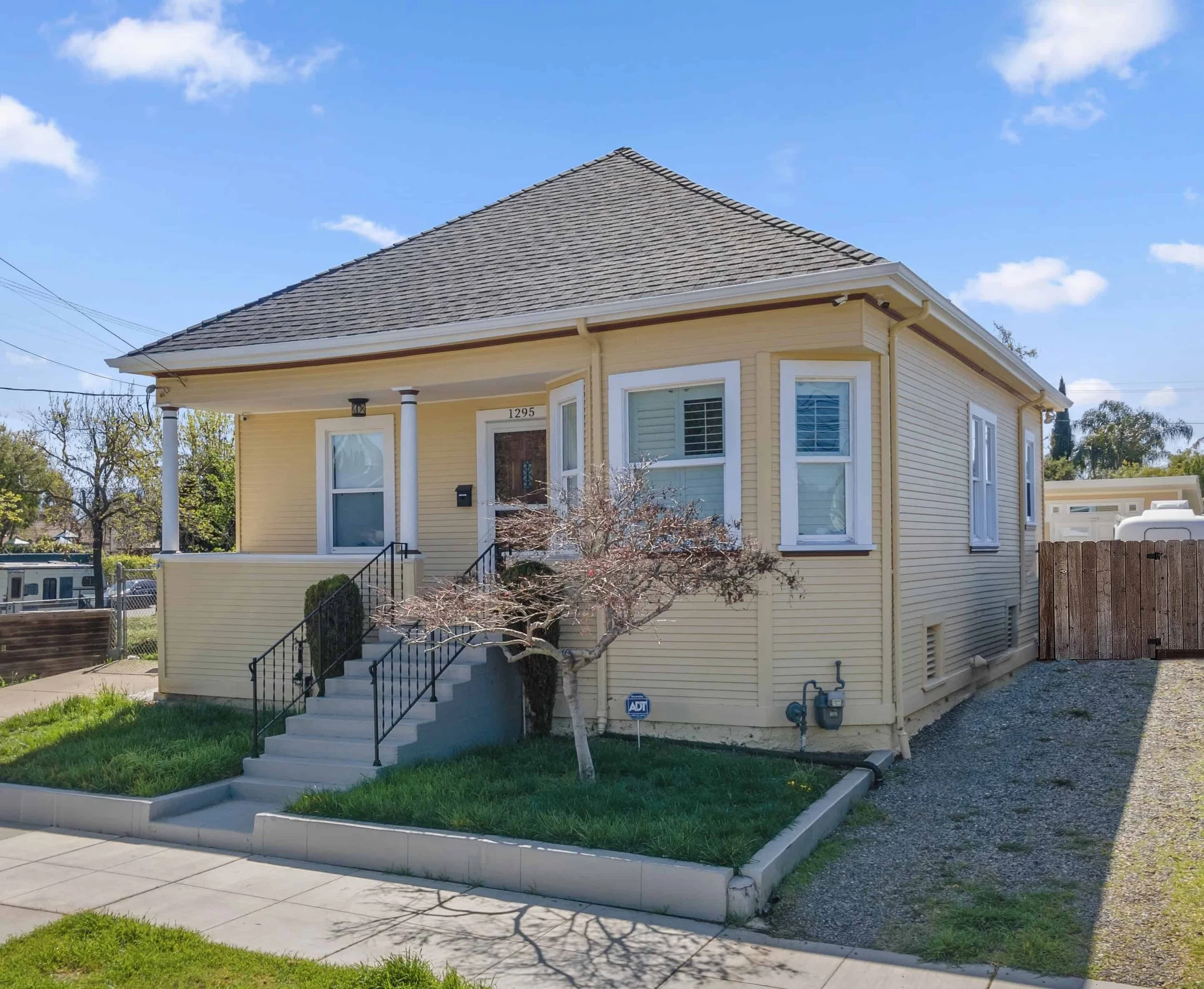 A yellow two-story house with a gabled roof and a small front porch with stairs, surrounded by a well-maintained lawn and a sidewalk. There are trees and a clear blue sky in the background.