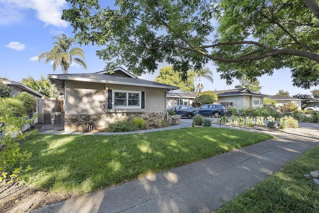 View of a suburban house with a well-maintained lawn, trees, and parked cars on the street, with bright sunlight and blue sky.