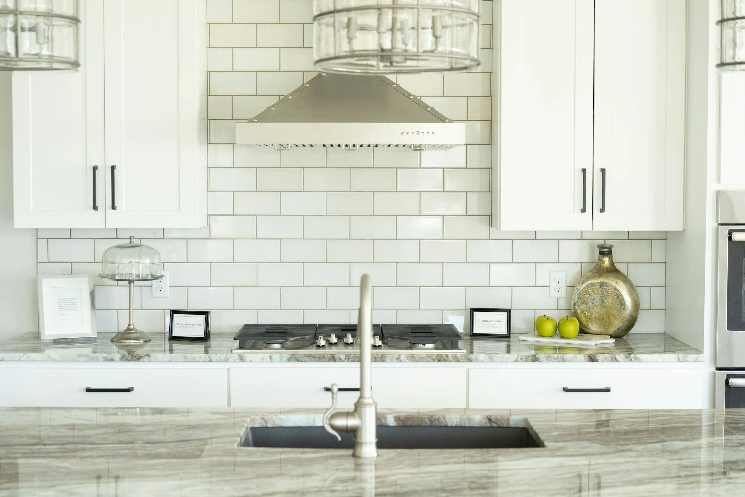 Modern kitchen with white cabinets, beige subway tile backsplash, stainless steel range hood, marble countertop, decorative objects including green apples, a glass cloche, and framed signs.