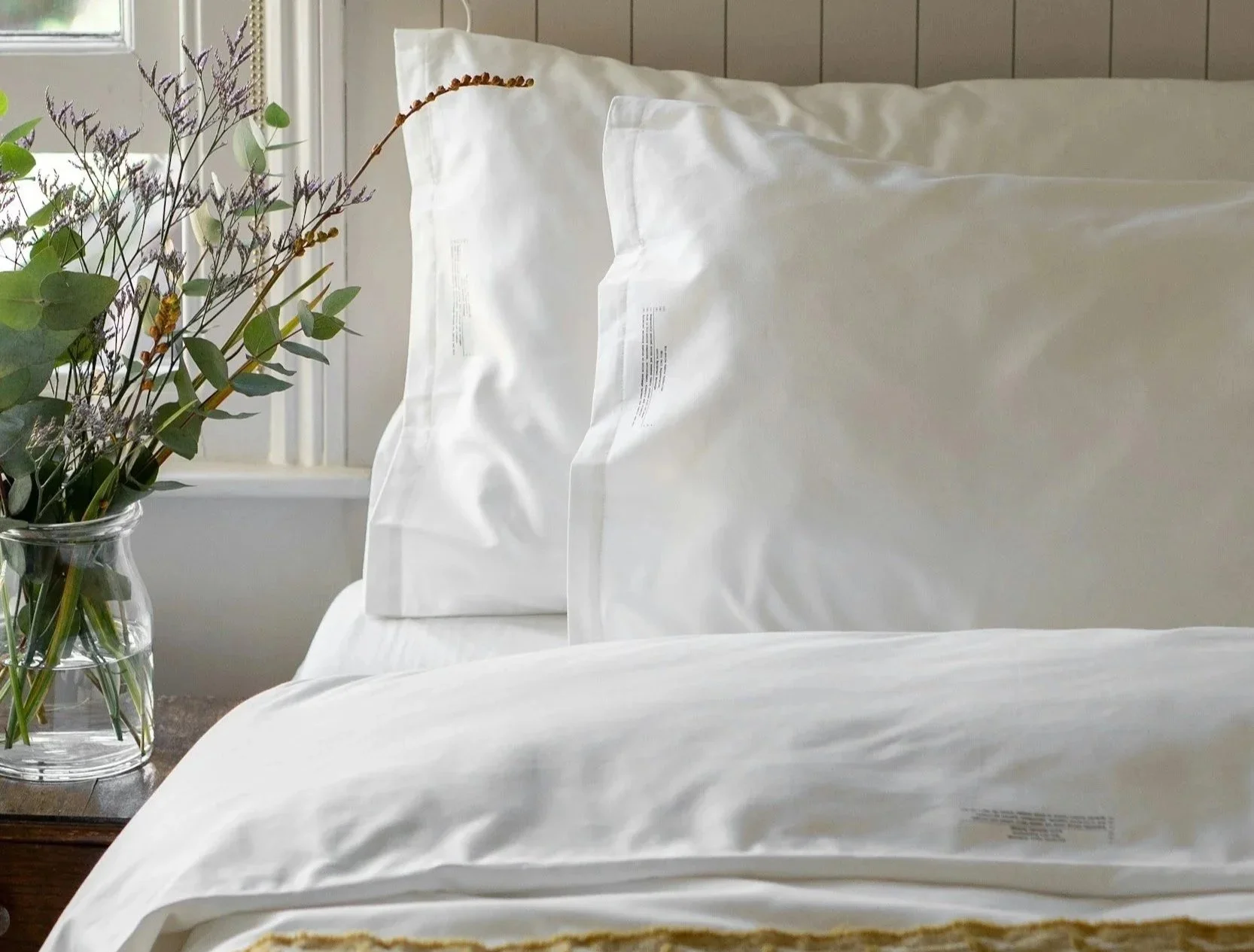 A bedroom with two white pillows on a bed with white sheets, a glass vase with flowers on a nightstand, and a window in the background.