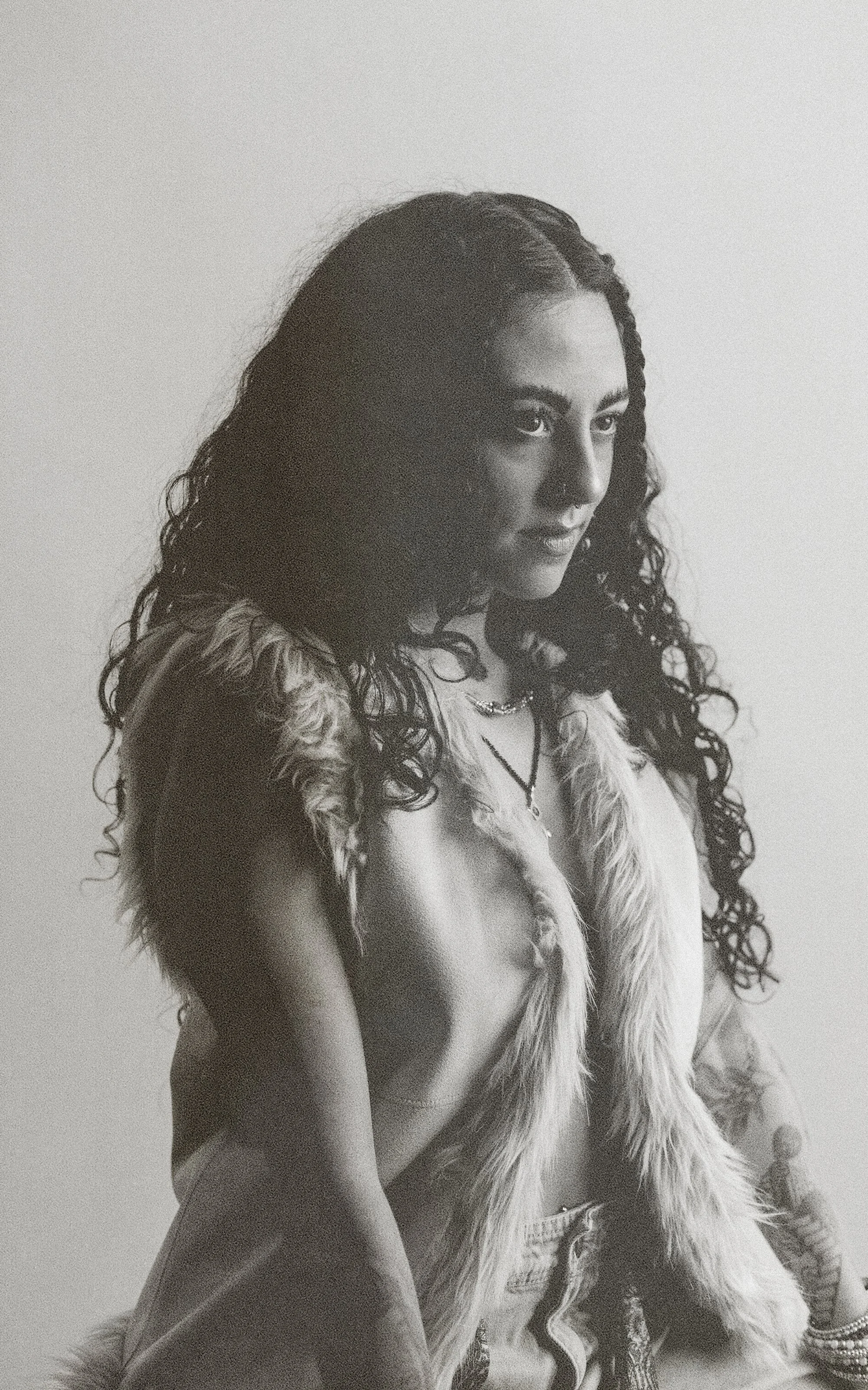 Black and white photo of a young woman with long curly hair, wearing a furry vest, layered necklaces, and bracelets, looking to the side with a serious expression.
