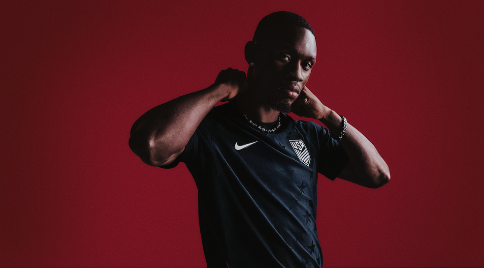A person with dark skin and short hair is adjusting a necklace against a red background, wearing a navy blue USA soccer jersey.