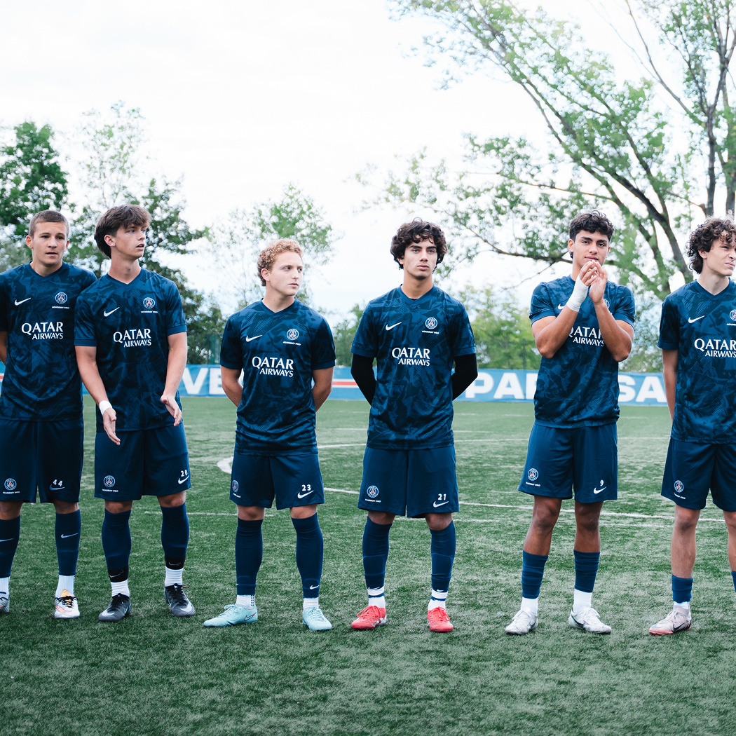 A group of young male soccer players standing on a field during the daytime. They are wearing navy blue uniforms with the PSG logo and Qatar Airways sponsorship. Some players are standing with their hands by their sides, while one has his hands clasped and another has their hands behind their back. Trees are visible in the background.