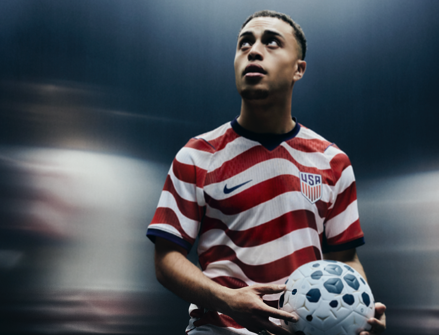 Young male soccer player in USA team jersey holding a soccer ball, looking upward against a gradient dark background.