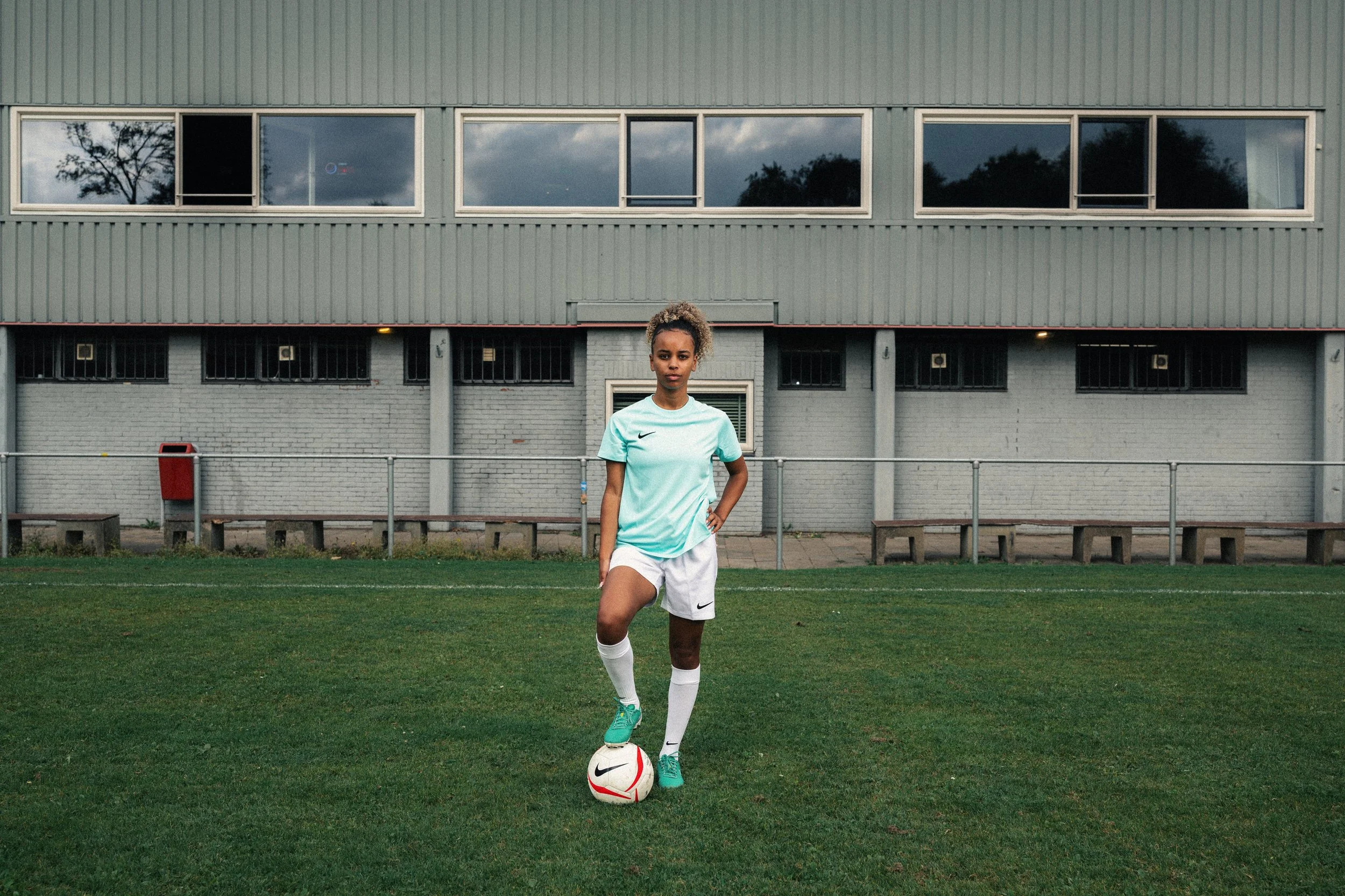 A female soccer player stands on a soccer field with her foot on a soccer ball, wearing a light blue jersey and white shorts, with a building and cloudy sky in the background.