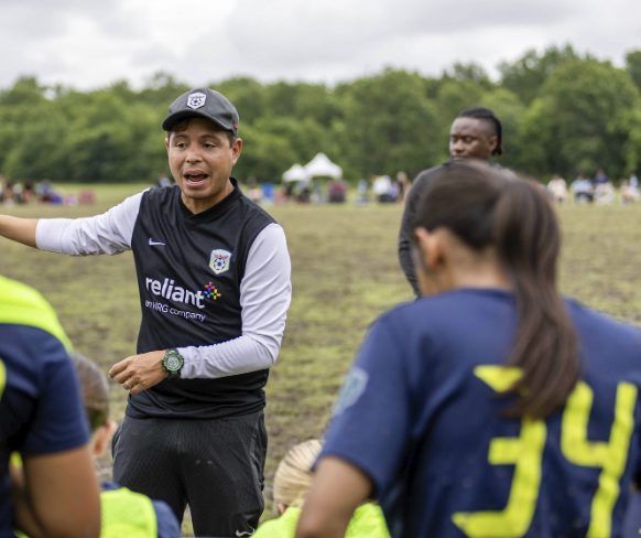 A coach in a black vest and gray long sleeve shirt, speaking to a team of female soccer players outdoors on a cloudy day.