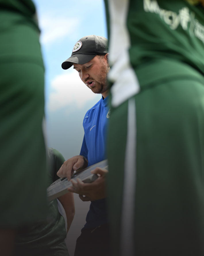 A man wearing a black baseball cap and a blue shirt, holding papers, seen through the gaps of two people wearing green sports jerseys.