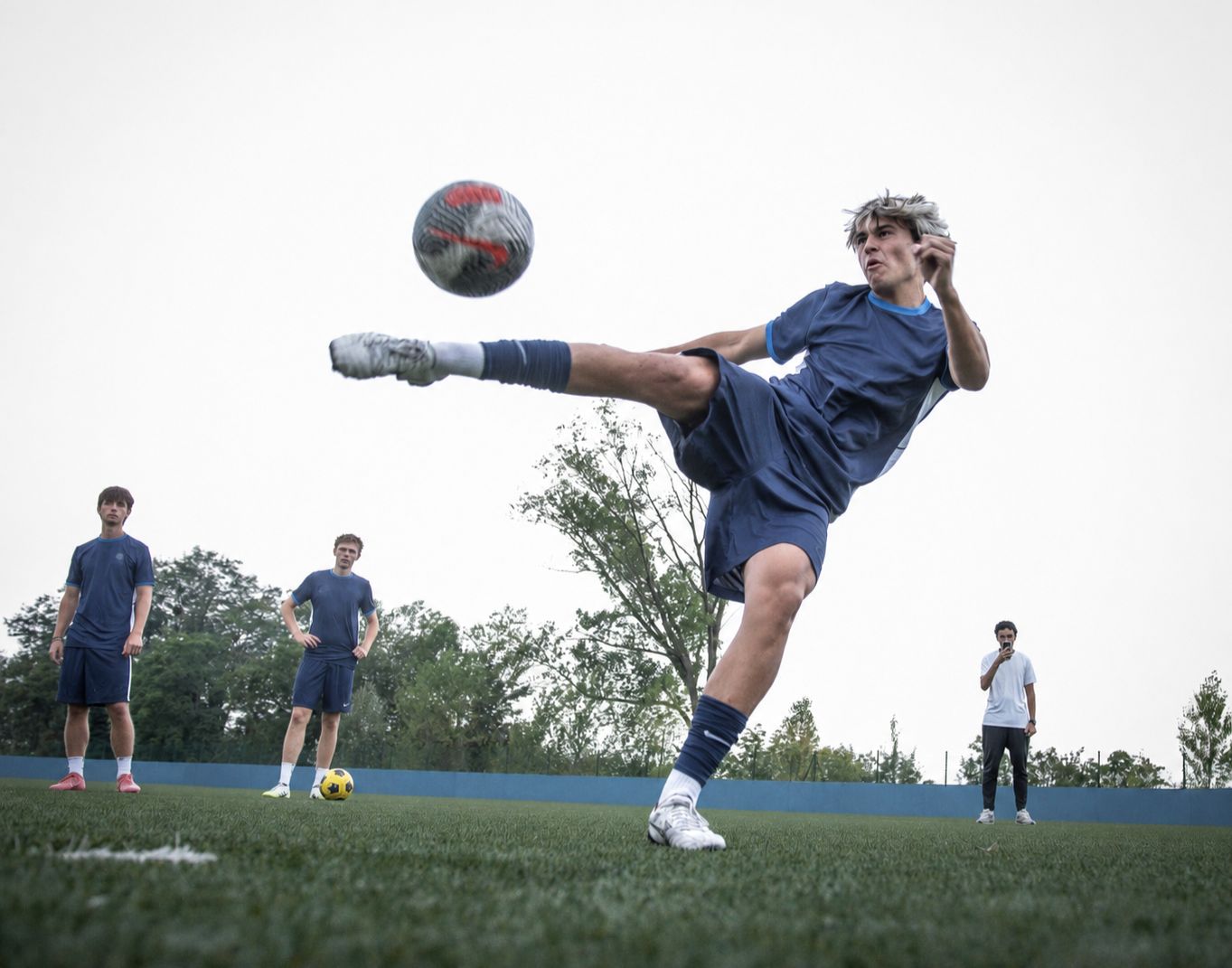 A soccer player in a blue uniform executes a high kick to strike the soccer ball during a match on a grassy field, with three other players and a person taking pictures in the background.