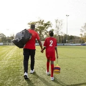 A soccer coach walking onto the field holding hands with a young player in a red uniform carrying a colorful bag.