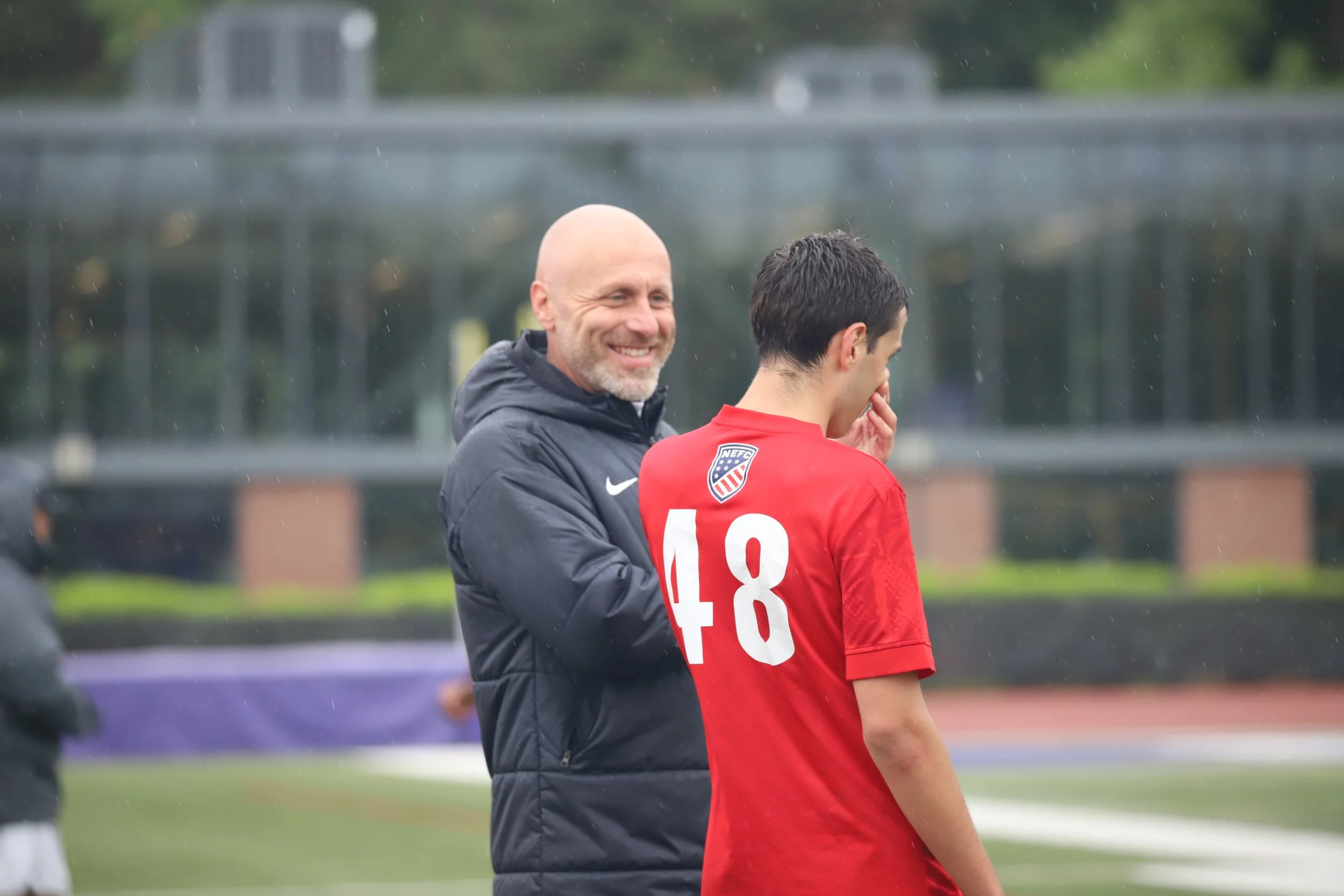 A soccer coach smiling and talking to a player in a red jersey with the number 18 on it during a game or practice on an outdoor field in rainy weather.