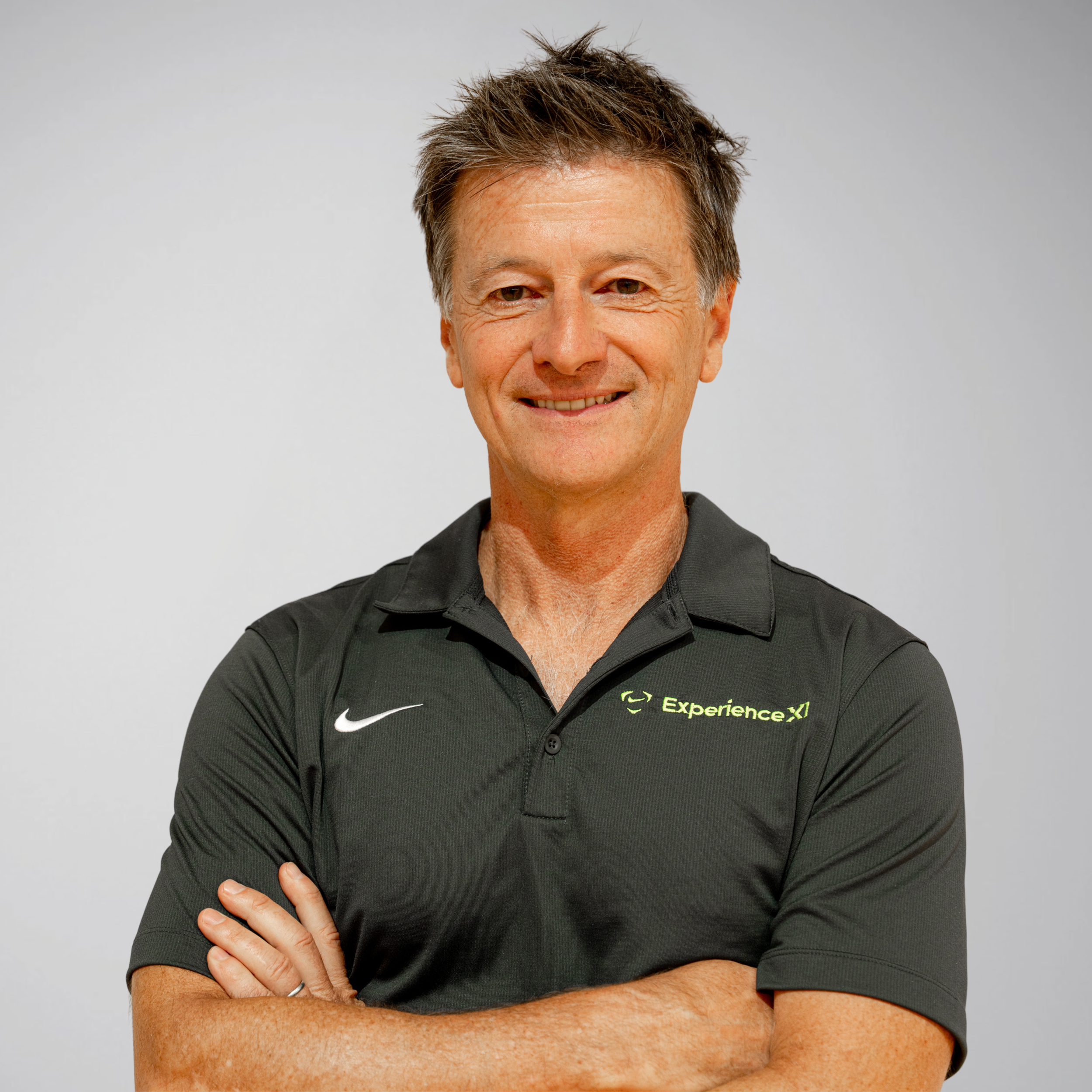 Close-up of a middle-aged man with short dark and gray hair, smiling, wearing a black polo shirt with the logo and text "ExperienceX" on it, standing against a plain light gray background.