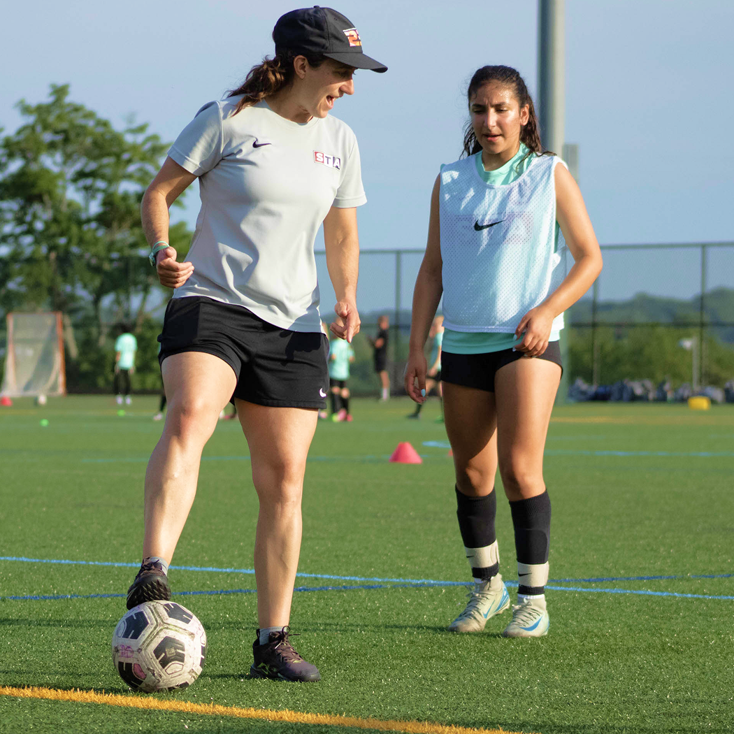 Two female soccer players practicing on the field, one is instructing the other.