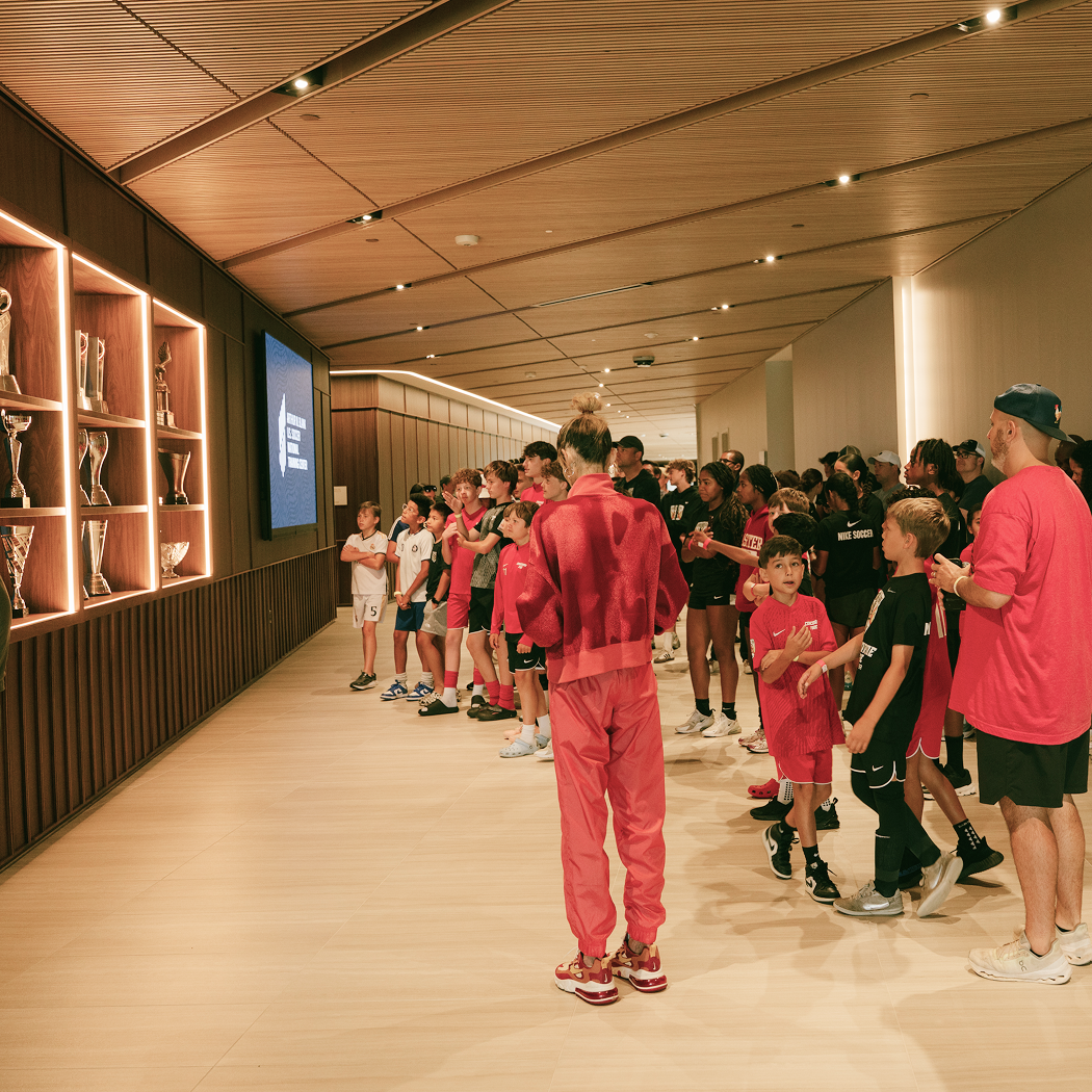 A group of children and adults standing in line inside a modern sports facility, looking at trophies displayed on a wall. Some children are wearing soccer jerseys, indicating a soccer event or practice.