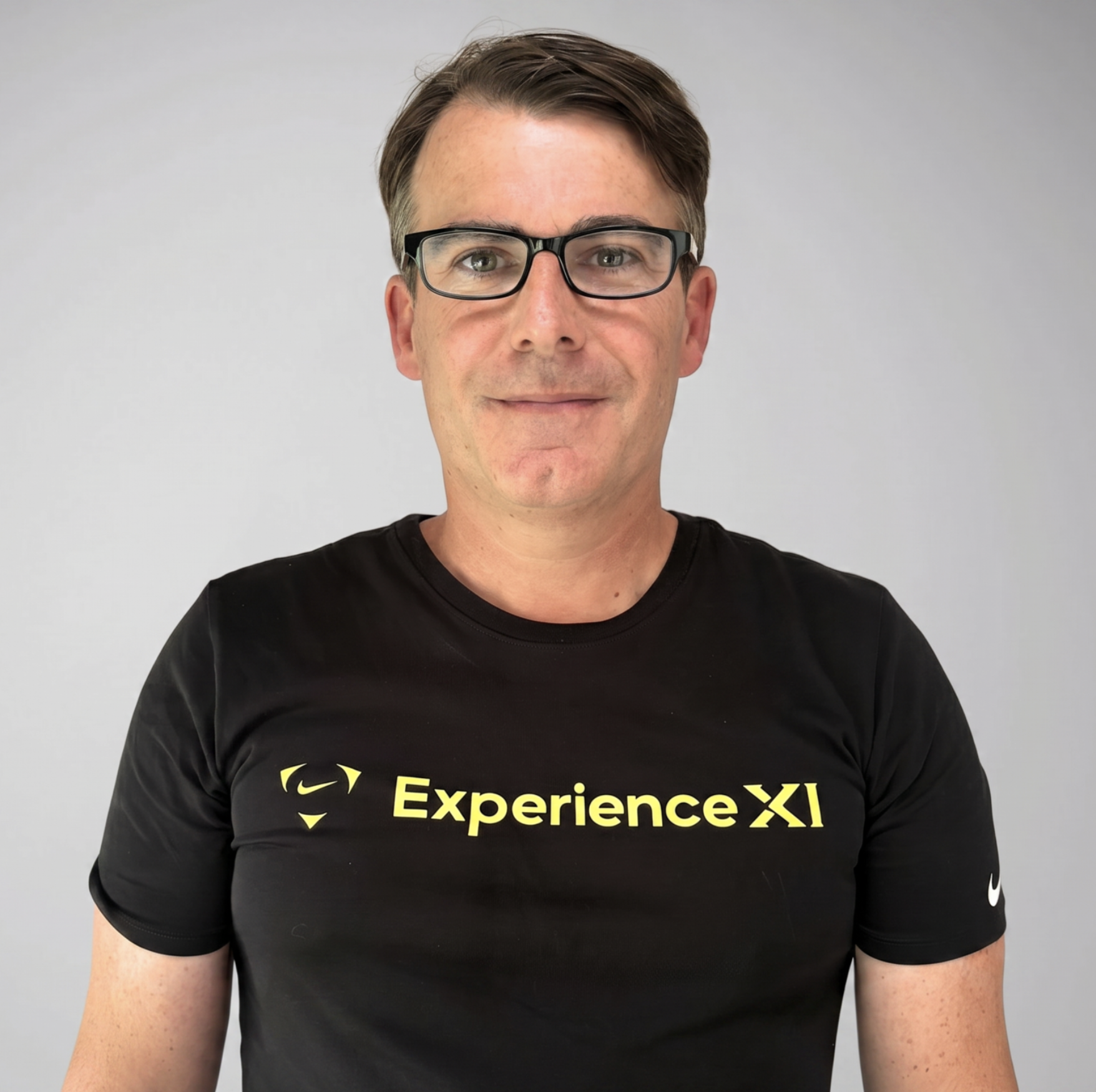 A man with short brown hair, glasses, and light skin, wearing a black T-shirt with yellow text that reads 'Experience XLI' and a yellow lion logo, standing against a plain gray background.