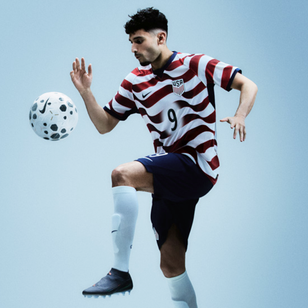 Male soccer player in USA jersey controlling a ball in mid-air against a light blue background.