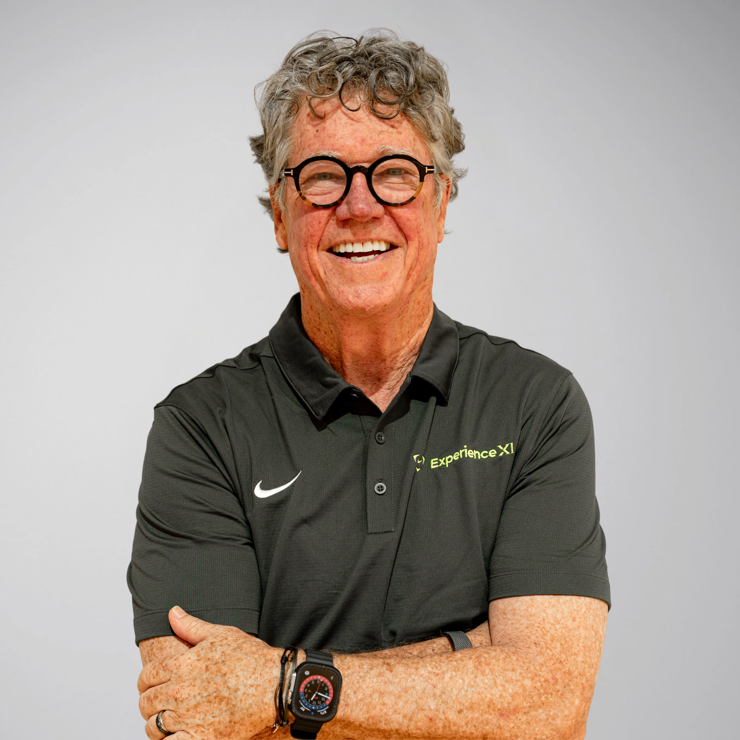A smiling elderly man with curly gray hair and glasses, wearing a black polo shirt, is standing with his arms crossed against a plain gray background.
