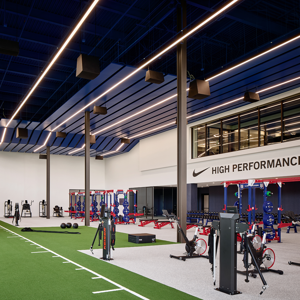 Modern gym with workout equipment, strength training stations, and cardio bikes, with a sign that reads 'High Performance' and football field markings on the floor.