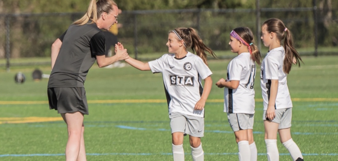A female coach or referee congratulating or encouraging a young girl on a soccer field during a game, with three other young girls waiting in line.
