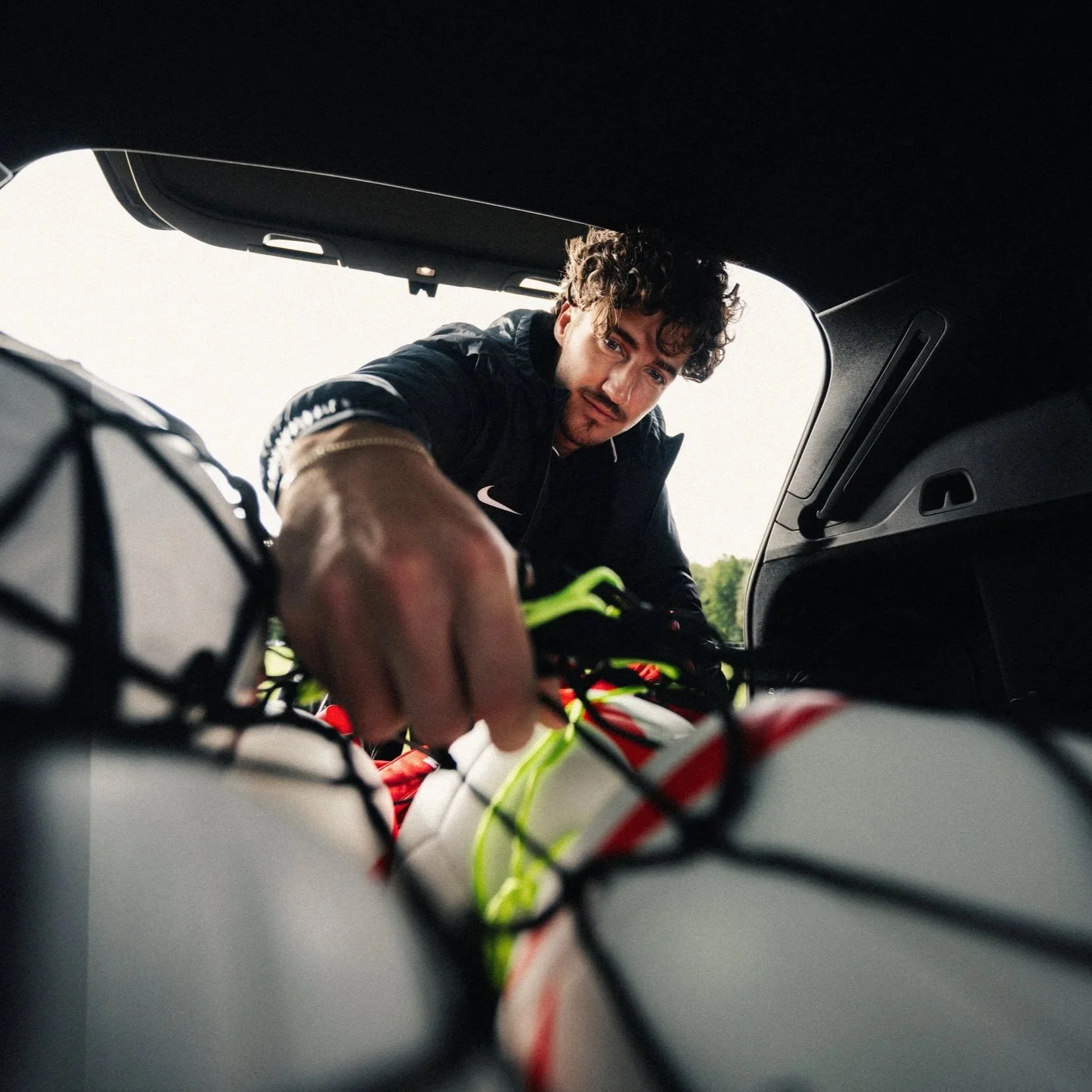 A man with curly hair is reaching into a car trunk, with a focus on a piece of equipment or gear with wires in the foreground. The scene appears to be outdoors.
