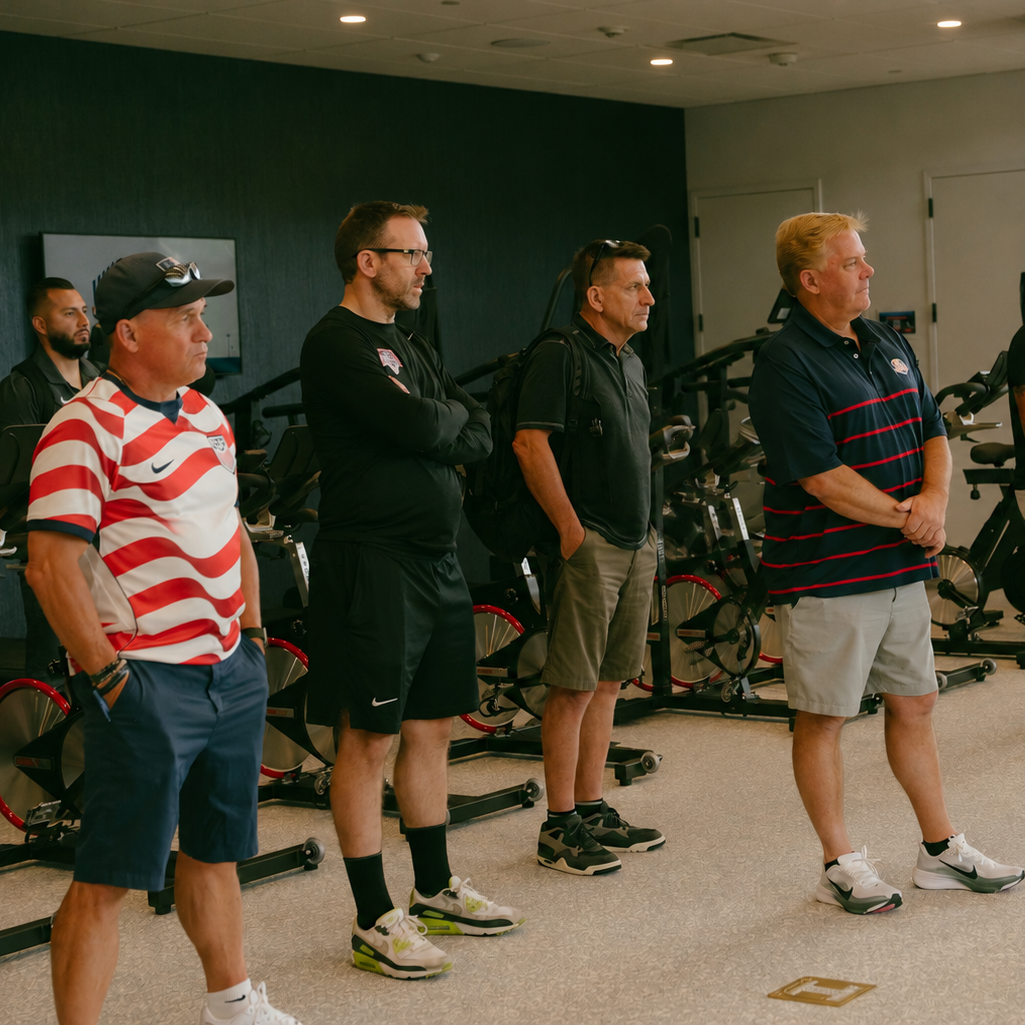 Four men standing in a gym, listening attentively. They are dressed in casual athletic clothing. Behind them are stationary bikes and exercise equipment.