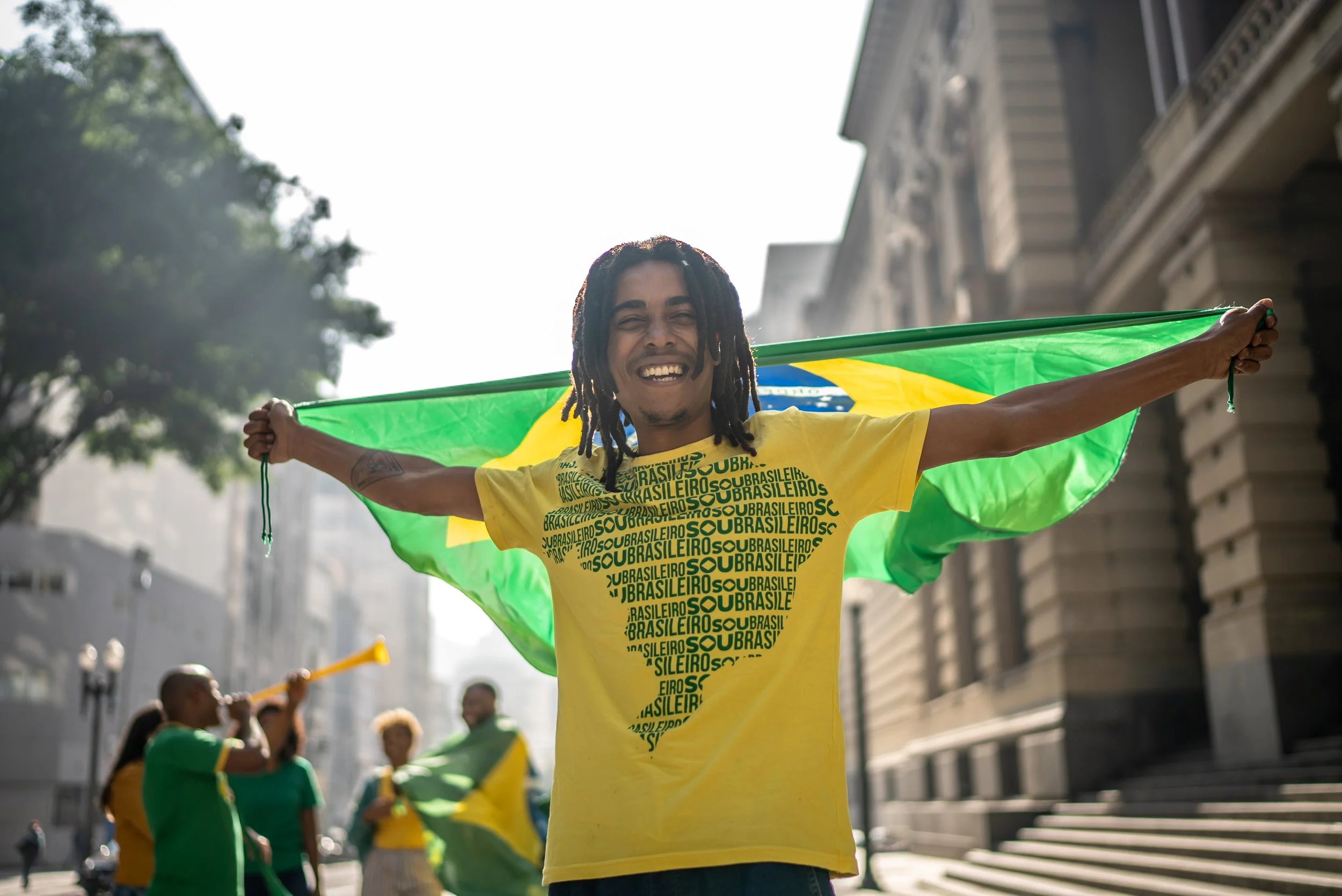 A man with dreadlocks smiling and holding a Brazilian flag behind his shoulders, standing in a city street with buildings and other people in the background.