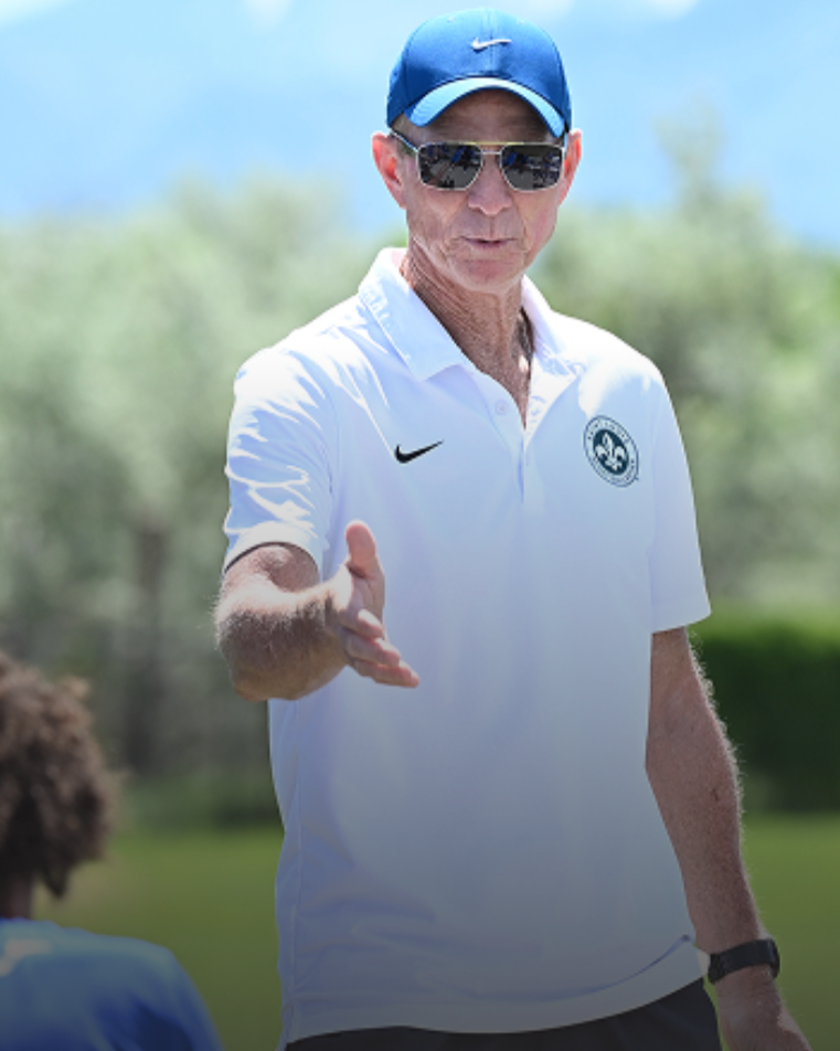 A man wearing a blue Nike cap, sunglasses, and a white Nike polo shirt with a logo, standing outdoors on a sunny day, extending his hand toward a person in a blue shirt.