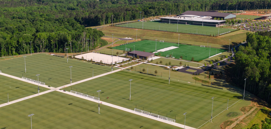 Aerial view of multiple sports fields, including soccer and football fields, surrounded by trees and parking lots.
