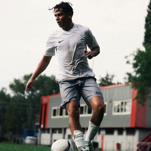 Young man playing soccer outdoors on a cloudy day, wearing a white sports shirt and gray shorts, kicking a soccer ball.