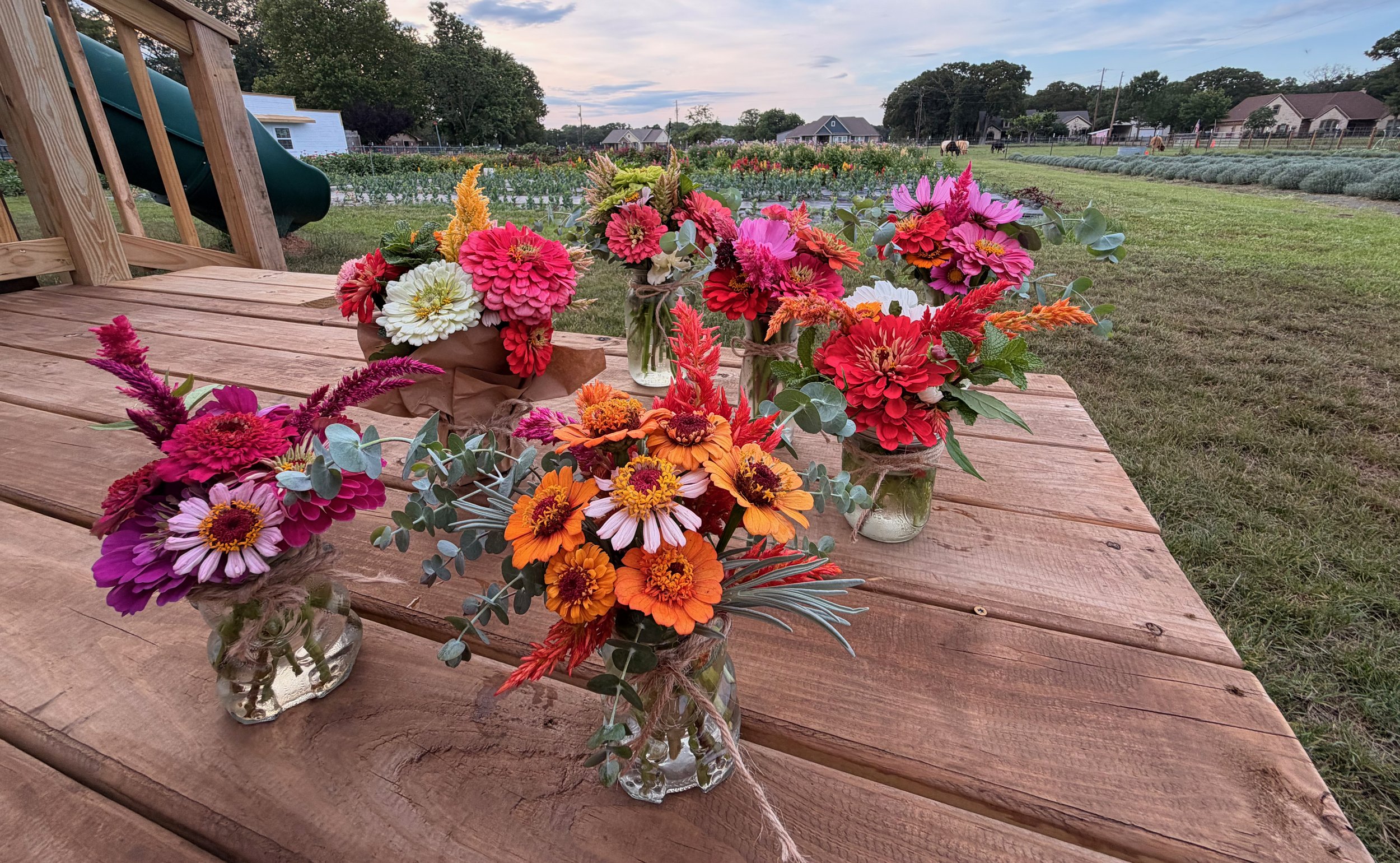 Multiple colorful flower bouquets in small glass jars on a wooden table outdoors, with a garden and houses in the background.