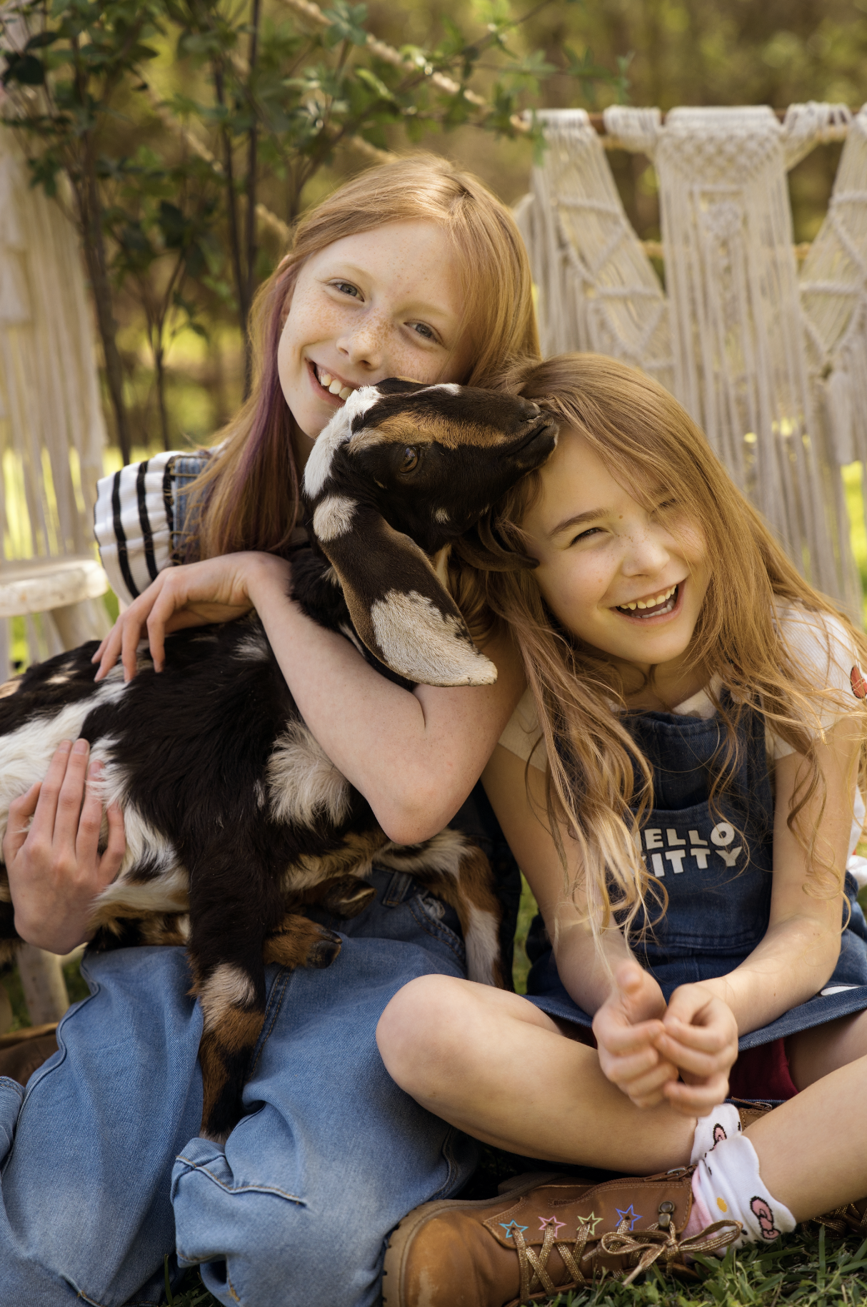 Two young red-haired girls smiling and laughing outdoors, hugging a black and white baby goat, with greenery and a white decorative fence in the background.