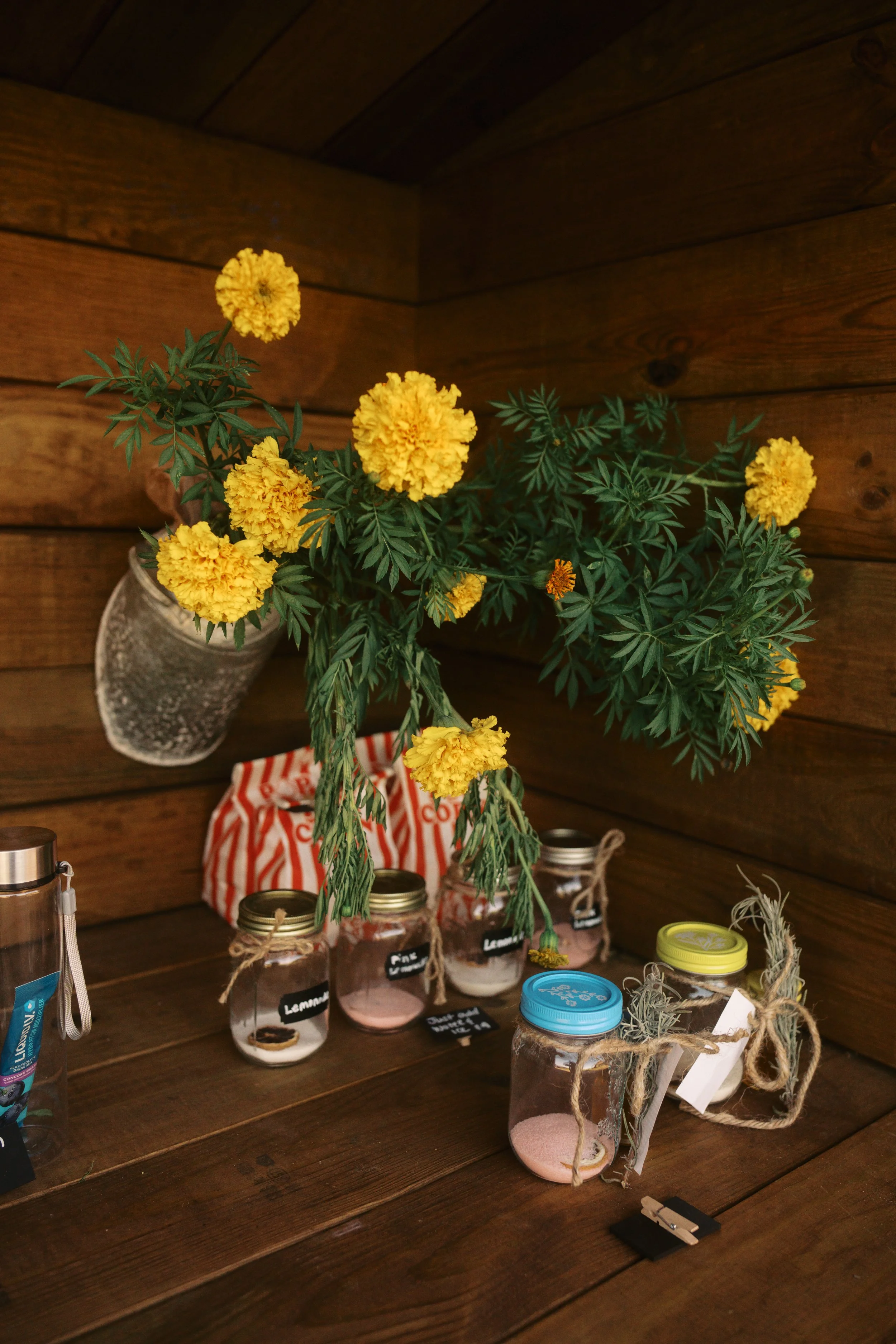 A wooden table with a yellow marigold flower arrangement in a white vase, several glass jars labeled with various herbs or spices, and jars with colorful lids, all set against a wooden wall.