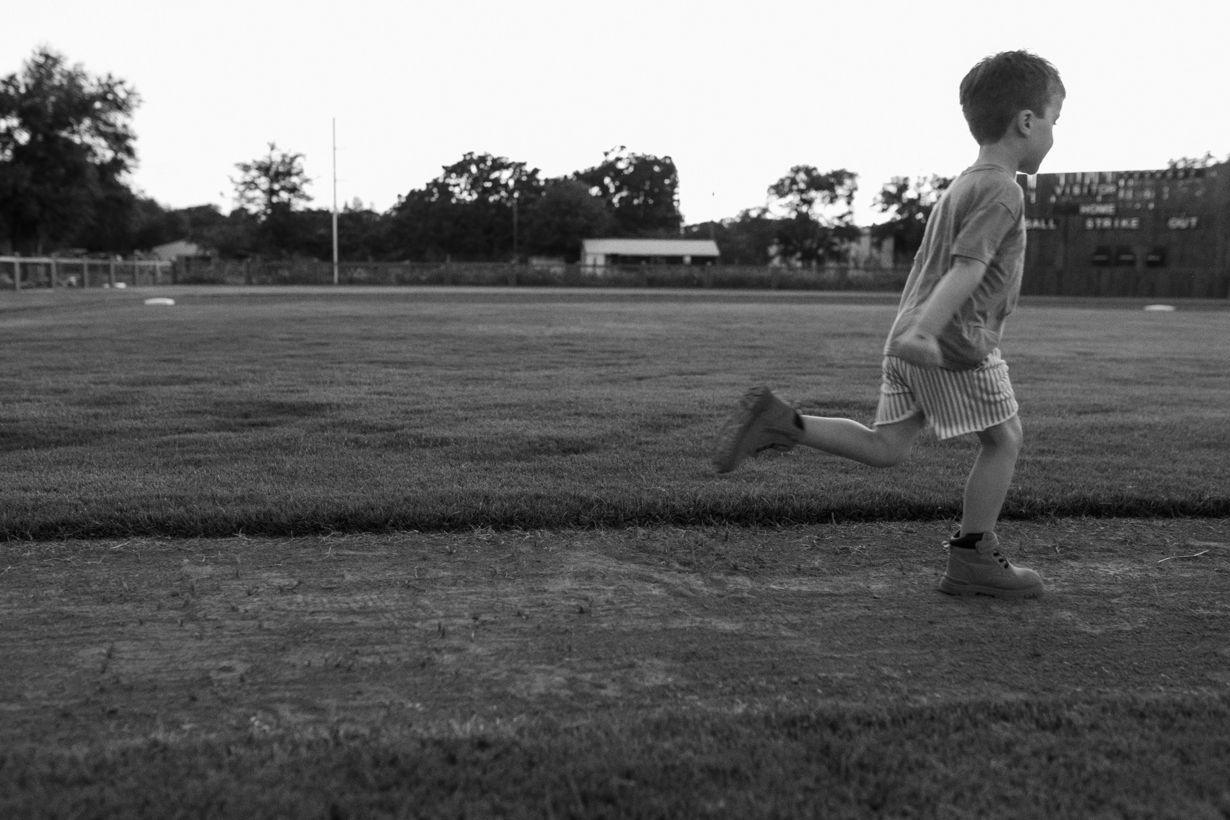 A young boy running on a grassy field at dusk, with trees and a building in the background.