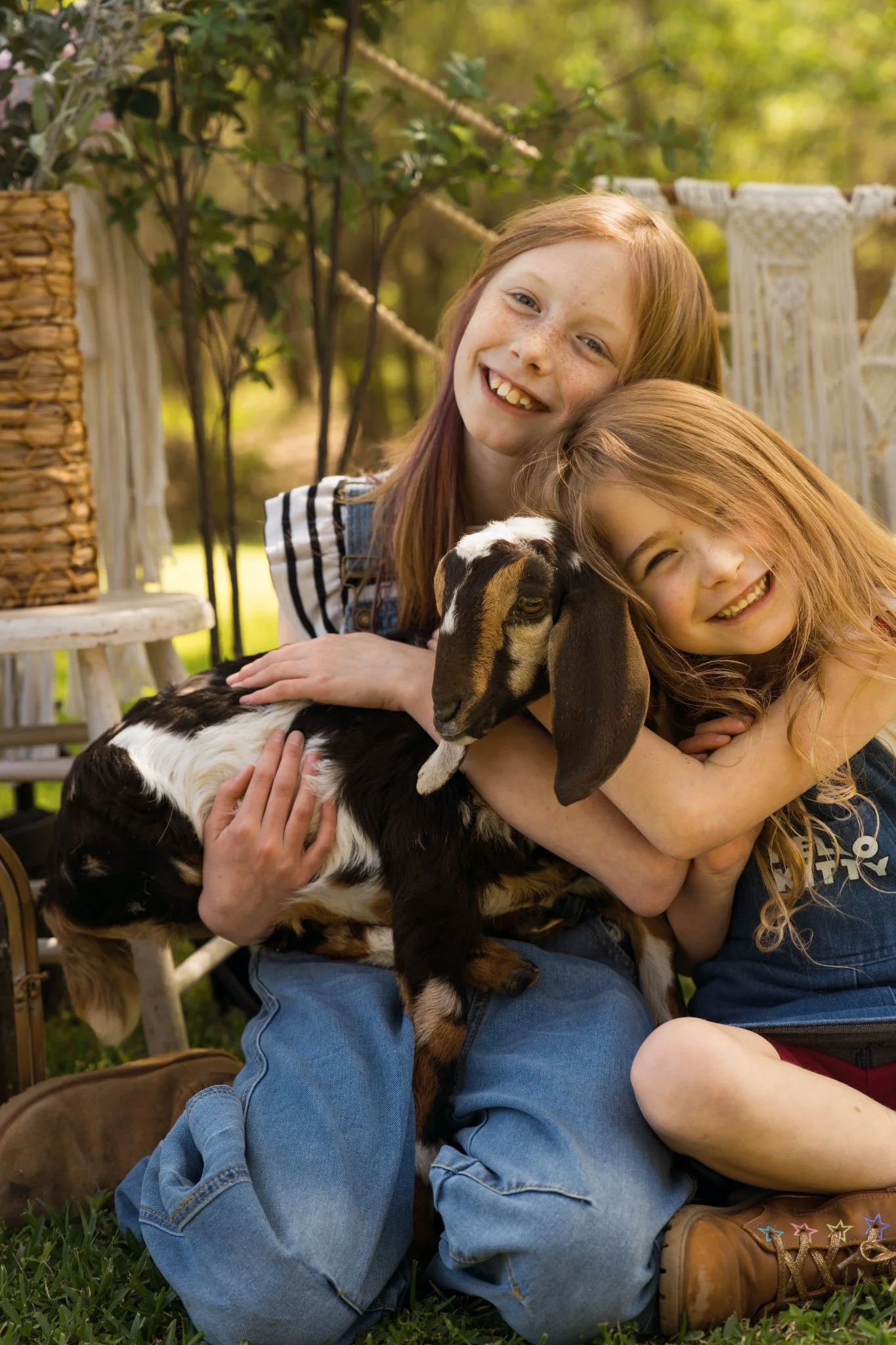 Two young girls hugging a black and white goat outdoors surrounded by green trees and grass.