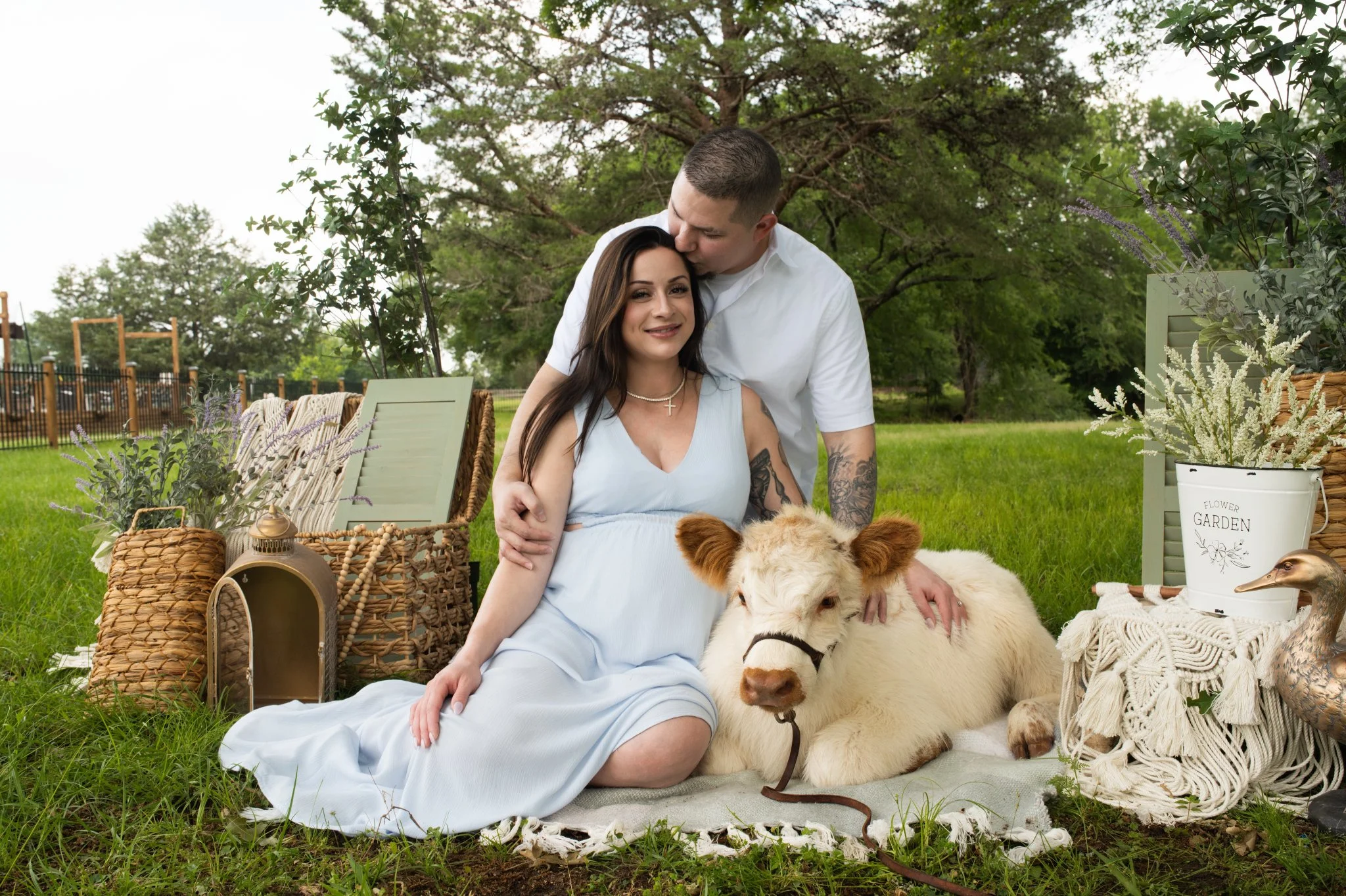 A pregnant woman sitting on a blanket outdoors, holding a white calf, with a man kissing her forehead. They are surrounded by decorative baskets, plants, and garden items in a green park with trees in the background.
