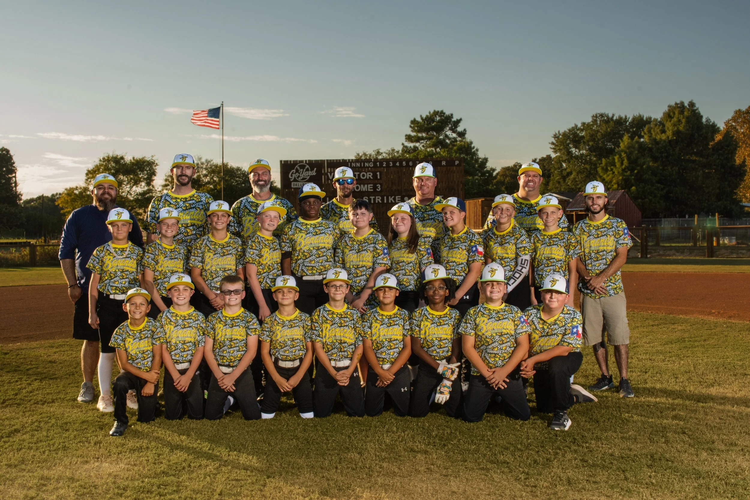 A group of children and coaches wearing baseball uniforms with yellow and blue designs, gathered on a baseball field for a team photo during sunset. The background shows trees, a scoreboard, and an American flag.