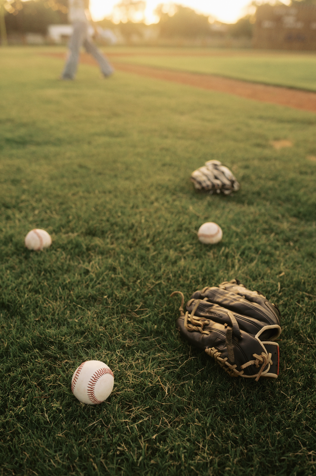 Baseball glove, baseballs, and baseball mitt on a grassy field during sunset, infield dirt visible in the background.