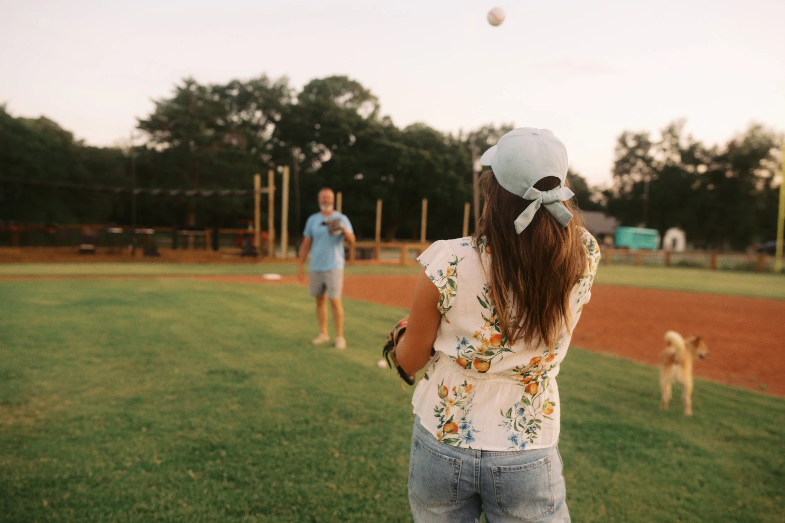 A girl wearing a white baseball cap with a bow, floral shirt, and denim shorts, playing baseball on a field. An older woman is in the background, holding a baseball glove, with trees and a baseball in the sky.