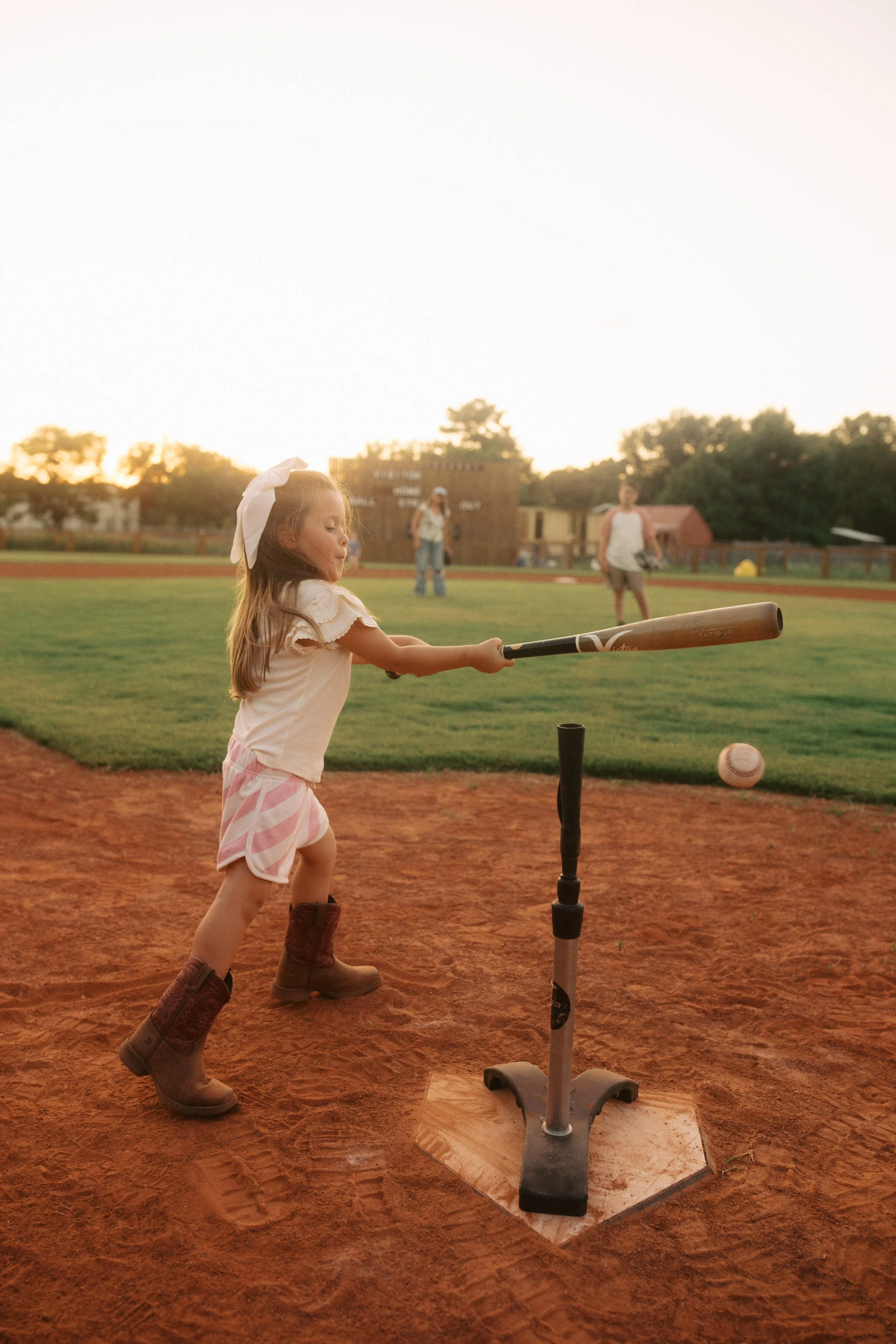 A young girl in a white shirt, pink and white striped shorts, and brown cowboy boots is swinging a baseball bat at a baseball being pitched towards her on a baseball field at sunset. There are a few people in the background and the sky is illuminated with the setting sun.